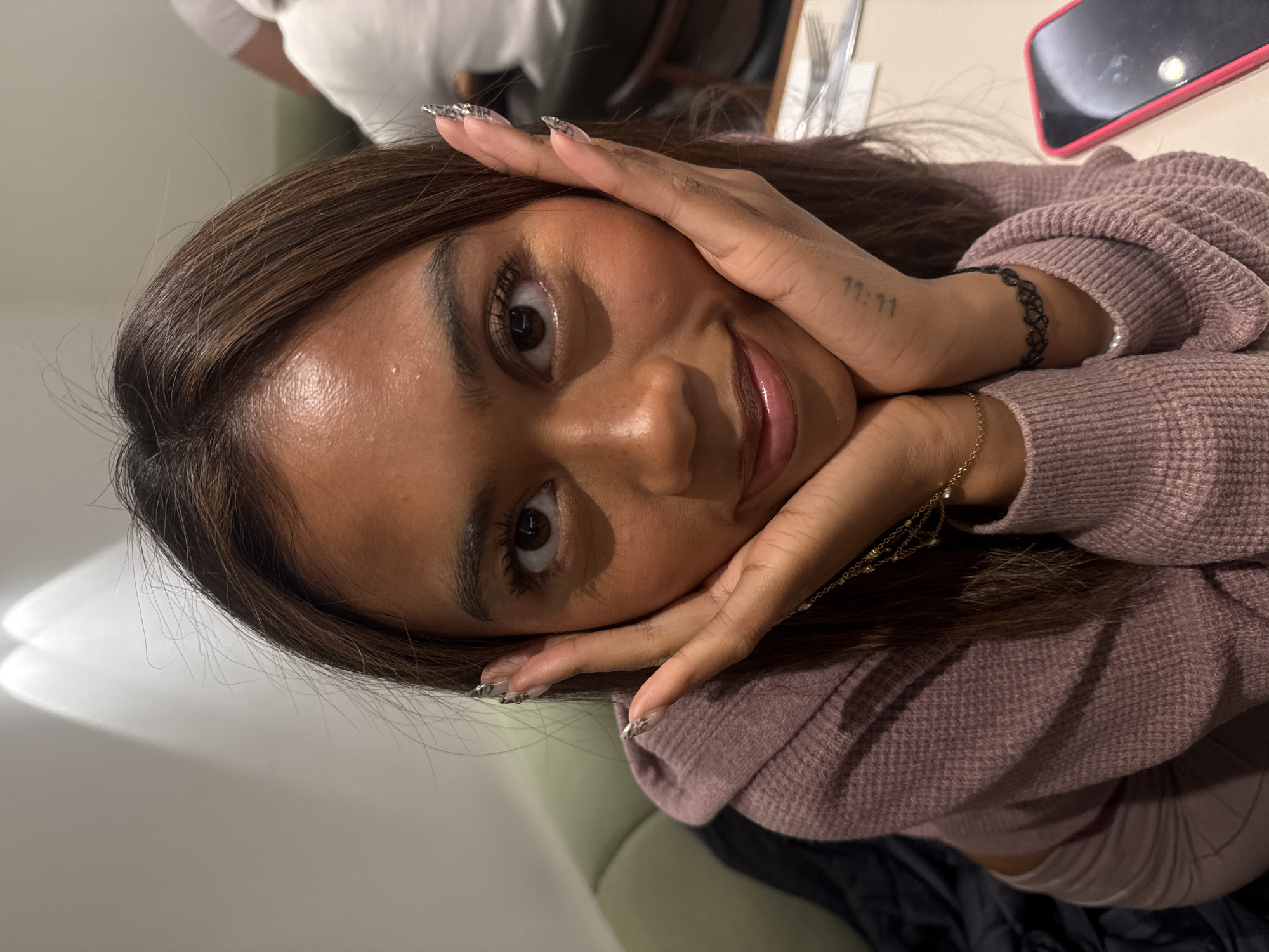 A young woman with tan skin and brown hair is centered in the image, her face close to the camera. Her hands are framing her face, and she is smiling. She is wearing a mauve sweater, a delicate gold necklace, and a black choker. A tattoo of "11:11" is visible on her wrist. Her nails are long and decorated with a white and silver design. In the background, a blurred table setting with cutlery and a red phone case can be seen. The environment suggests an indoor setting, possibly a restaurant. The location in the city of London, United Kingdom, cannot be confirmed.