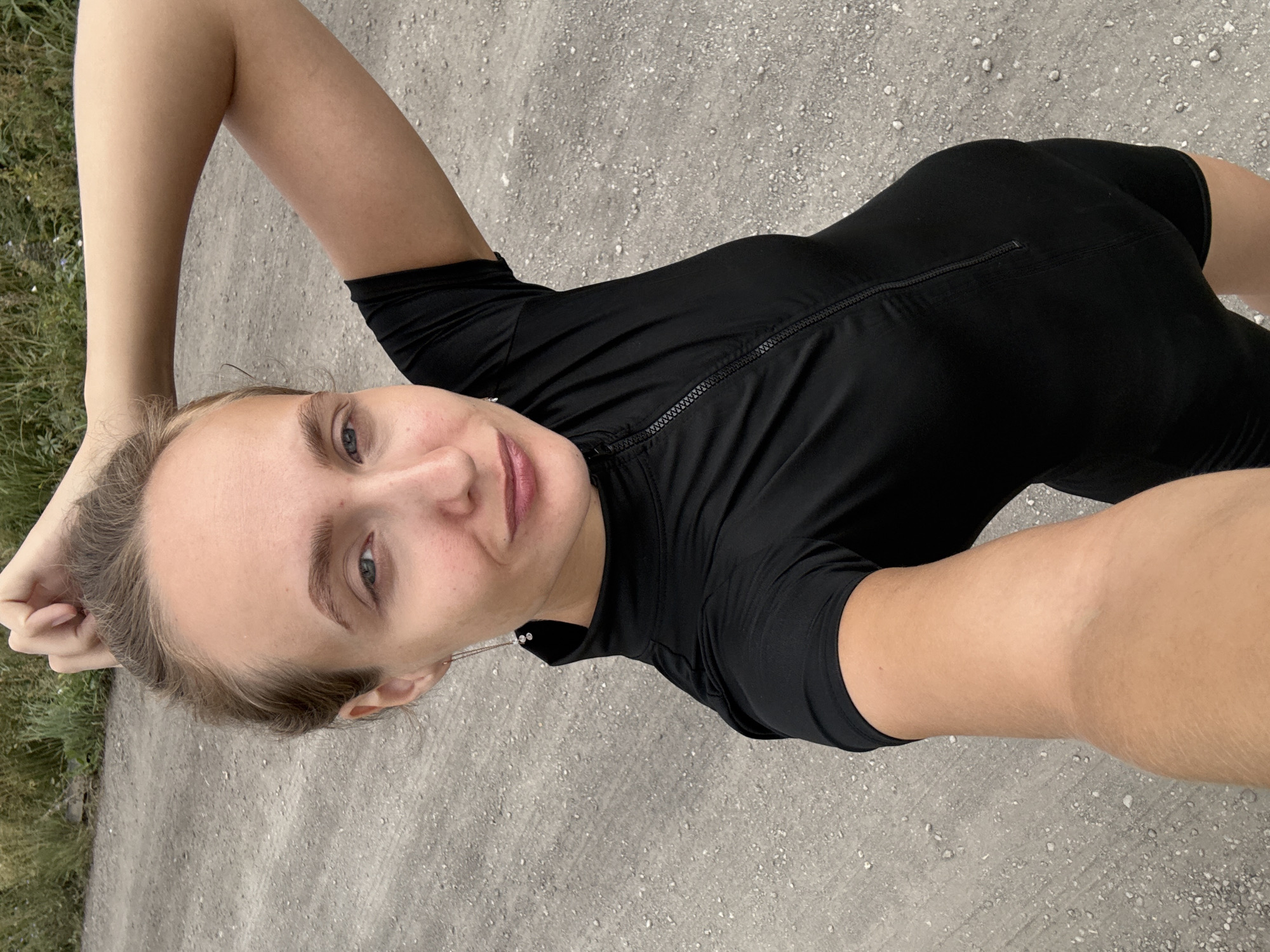 A woman is taking a selfie outdoors. She is wearing a black short-sleeved, high-necked garment with a zipper down the front. Her right arm is raised and bent, with her hand resting on the back of her head. She has light-colored hair pulled back and is looking at the camera. She is wearing small, dangling earrings. The background appears to be a dirt or gravel road with some dry grass and vegetation to the left. The scene suggests an outdoor, possibly rural or semi-rural, environment, potentially in or near the village of Podshivalovo, Russia, given the context.