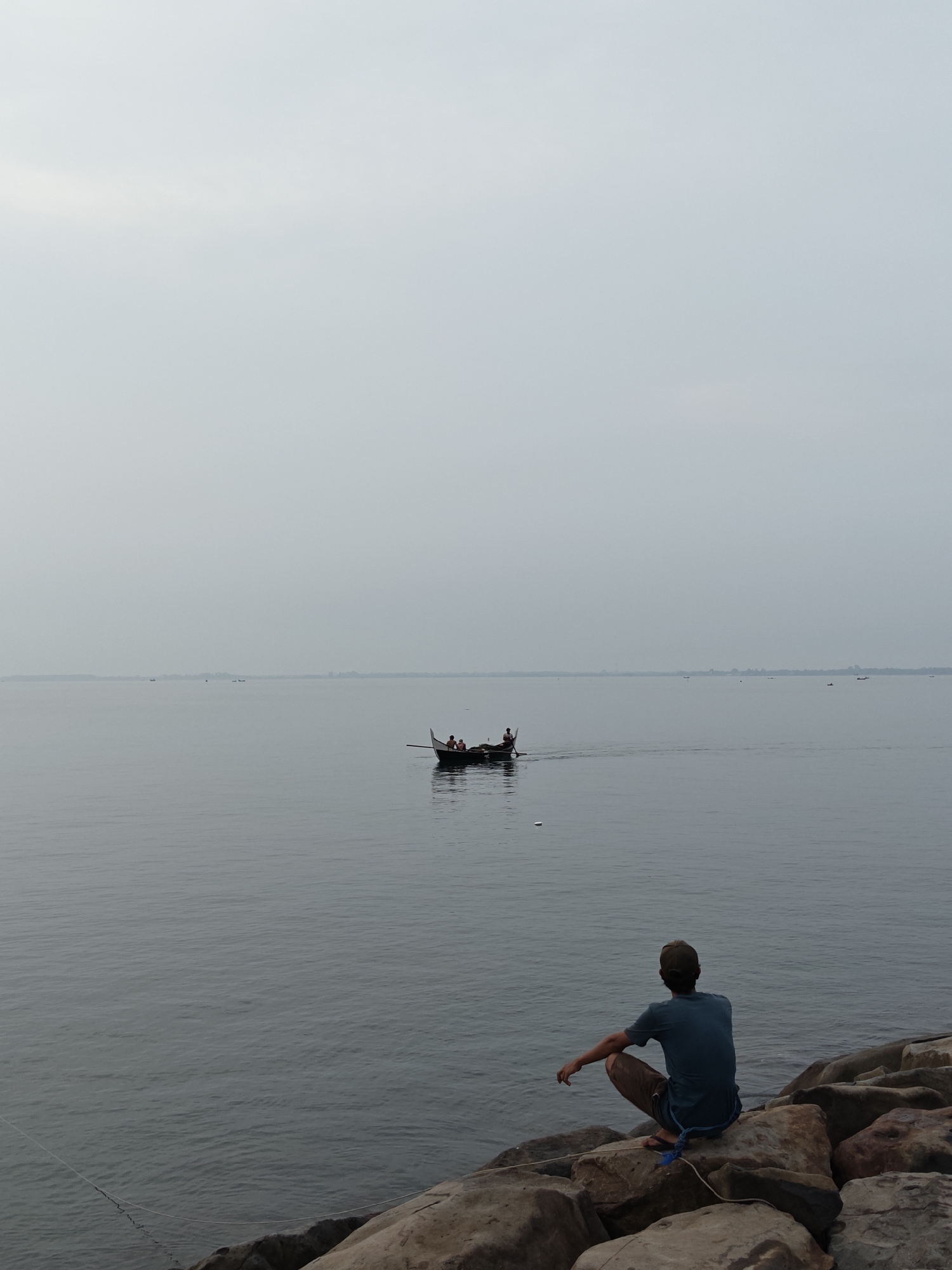 The image captures a serene waterside scene, likely in Lhokseumawe, Indonesia, under an overcast sky. In the foreground, a person is seen from behind, sitting on large, weathered rocks that form a natural barrier along the shore. The individual wears a blue short-sleeved shirt, brown pants, and a cap, and is looking out across the calm, grey water. A blue rope or line is visible near their feet, extending towards the water, suggesting potential fishing or some other waterside activity.
The mid-ground features a small, traditional wooden boat with two individuals aboard. One person appears to be paddling or operating the boat, which leaves a subtle wake behind it as it moves across the smooth surface of the water. In the distant background, several other faint, small boats can be seen scattered across the wide expanse of water, with a low, indistinct landmass or horizon line barely visible beneath the very light, almost white, sky. The overall atmosphere is quiet and contemplative, characterized by muted tones of grey, blue-grey, and brown. There is no visible text in the image.