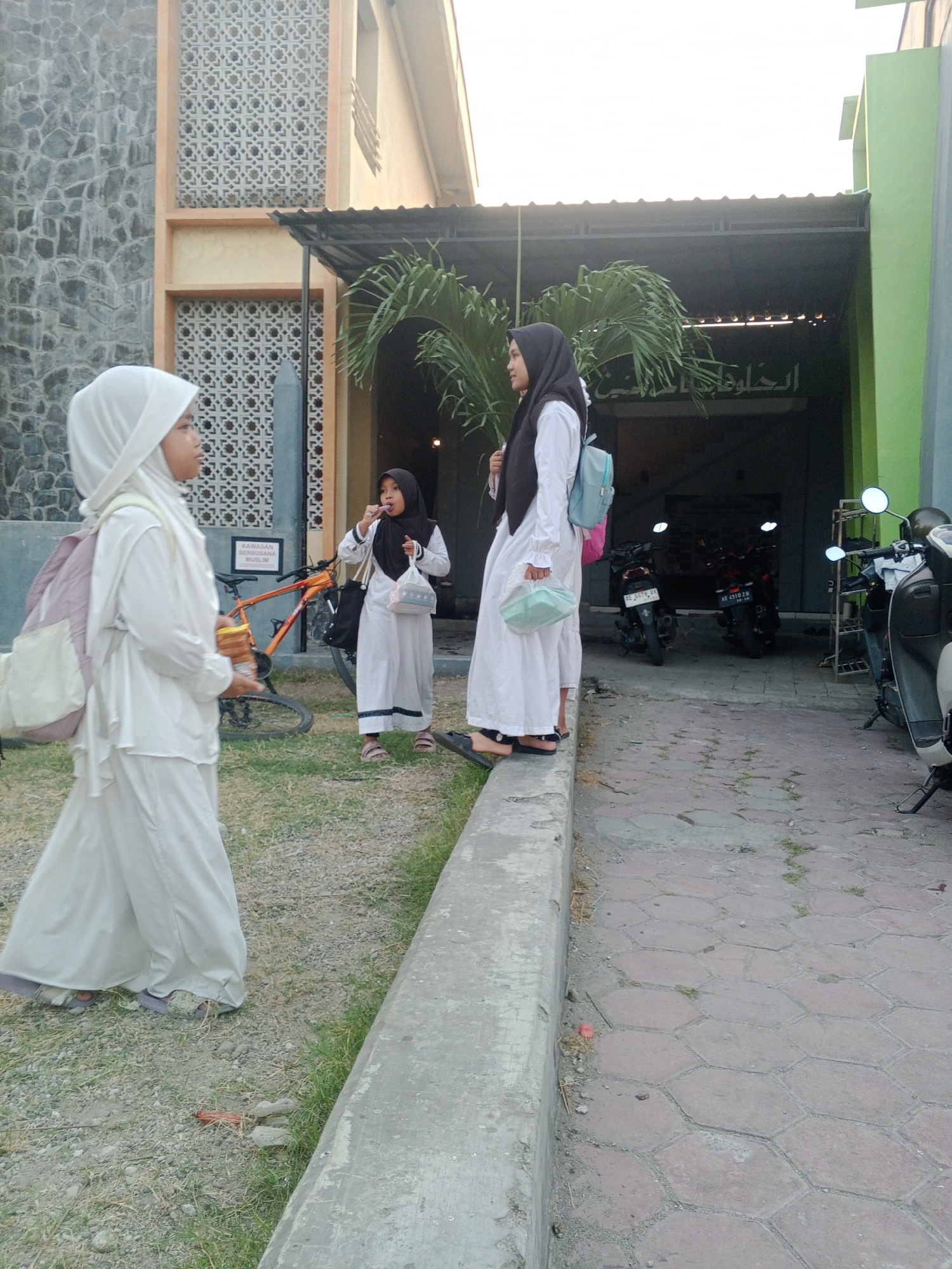 The image depicts an outdoor scene near a building, possibly in Lembah, Indonesia. Three young girls wearing white Islamic attire and headscarves are present. The girl on the left is walking away from the camera, carrying a light pink backpack. The middle girl is standing, holding a light blue bag and appears to be eating something from a small white package. The girl on the right is also standing and holding a light blue bag. Behind the girls, there is a building with stone and patterned wall details, a roof, and some greenery. A bicycle is visible in the background, along with several motorcycles parked under the building's overhang. The ground is a mix of gravel and paved hexagonal tiles.