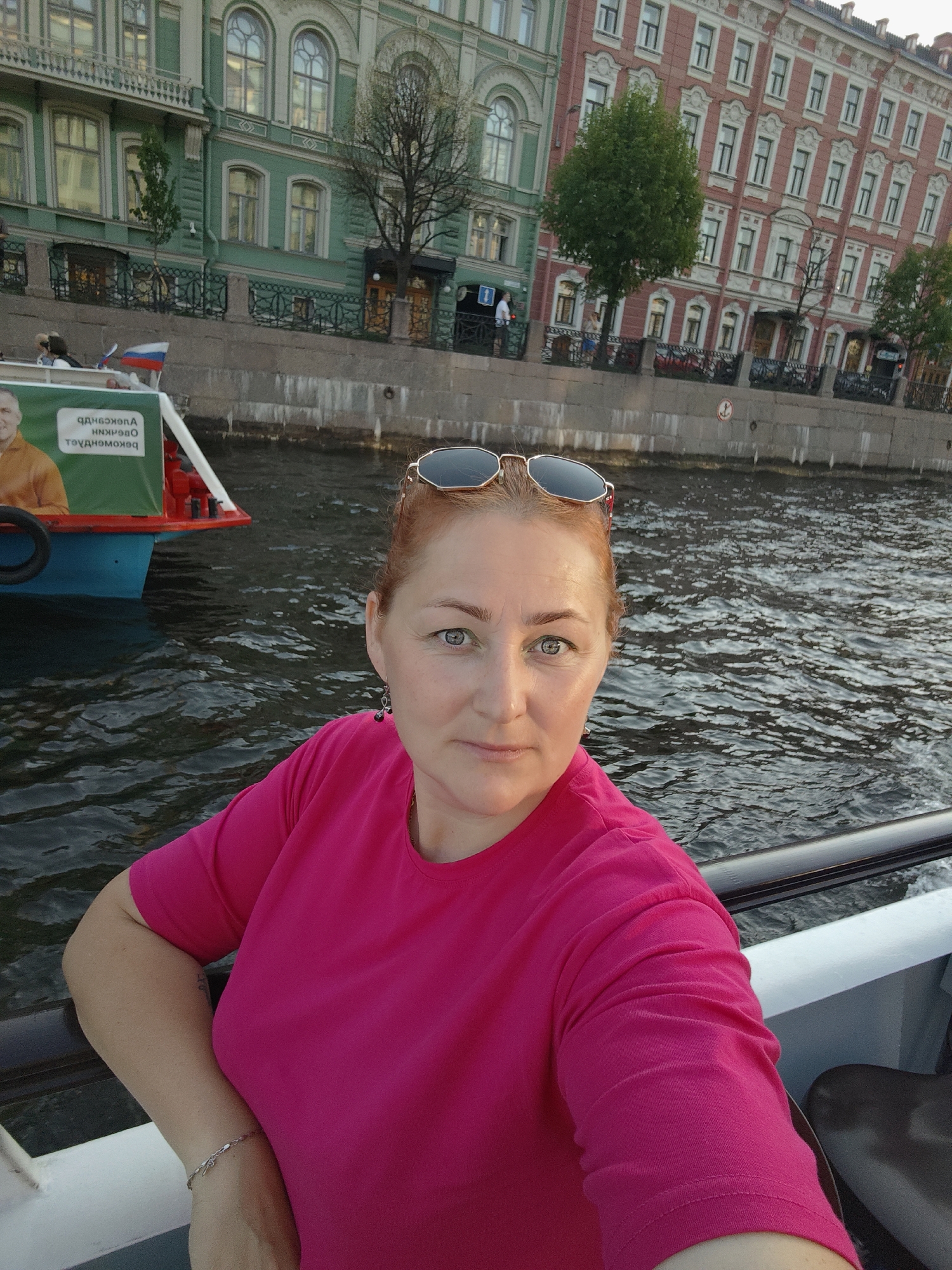 A woman with auburn hair, wearing a bright pink t-shirt and sunglasses, is taking a selfie from a boat. She is on the water with buildings in the background. The buildings appear to be historic and are located along a canal or river. The larger, red building on the right is likely in the neoclassical style, common in St. Petersburg, Russia. The green building on the left also exhibits architectural features typical of the city. The water is rippling, suggesting the boat is moving. A portion of the boat's railing and a seat are visible in the foreground. A green banner with Cyrillic text and a picture of a man is attached to the boat on the left side. A Russian flag is also visible on the boat. The overall scene suggests a tourist activity on a waterway in St. Petersburg, possibly during a guided tour of the city's famous canals.