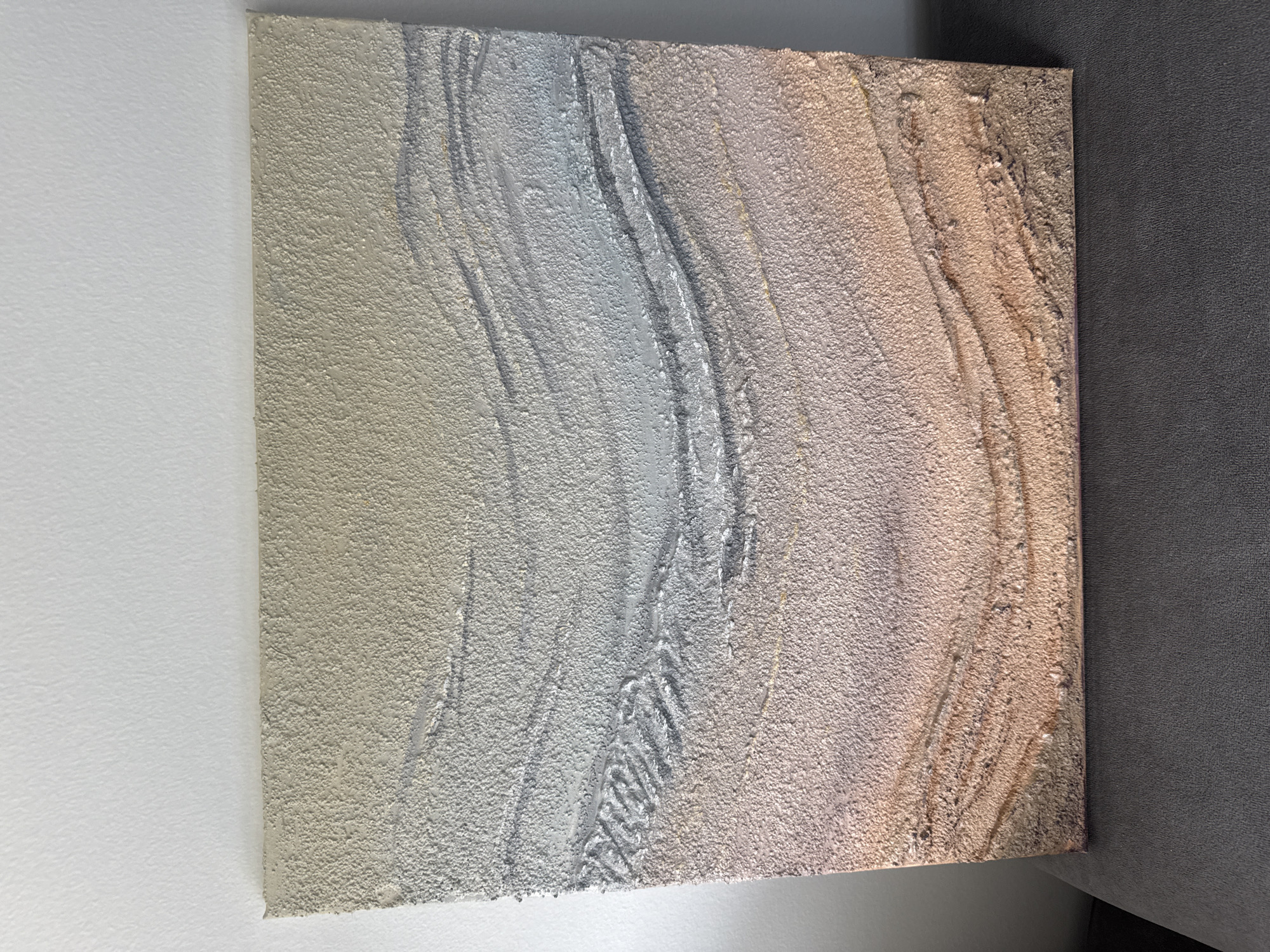 The image shows a textured abstract painting, leaning against a white wall. The painting is square and features wavy, layered bands of color. The top band is a pale beige, transitioning to a silver-grey middle section with darker grey lines. The bottom section is a gradient of pale pink to sandy brown, with some darker brown and gold accents. The texture of the painting appears rough and granular, with raised ridges and indentations that create shadows. The painting is displayed indoors, with the lower portion resting on a dark grey upholstered surface, possibly a sofa or chair. The lighting suggests a natural light source from the left, casting subtle shadows on the painting and the wall behind it. There is no indication of the city of Limassol, Cyprus, or any specific location within it.