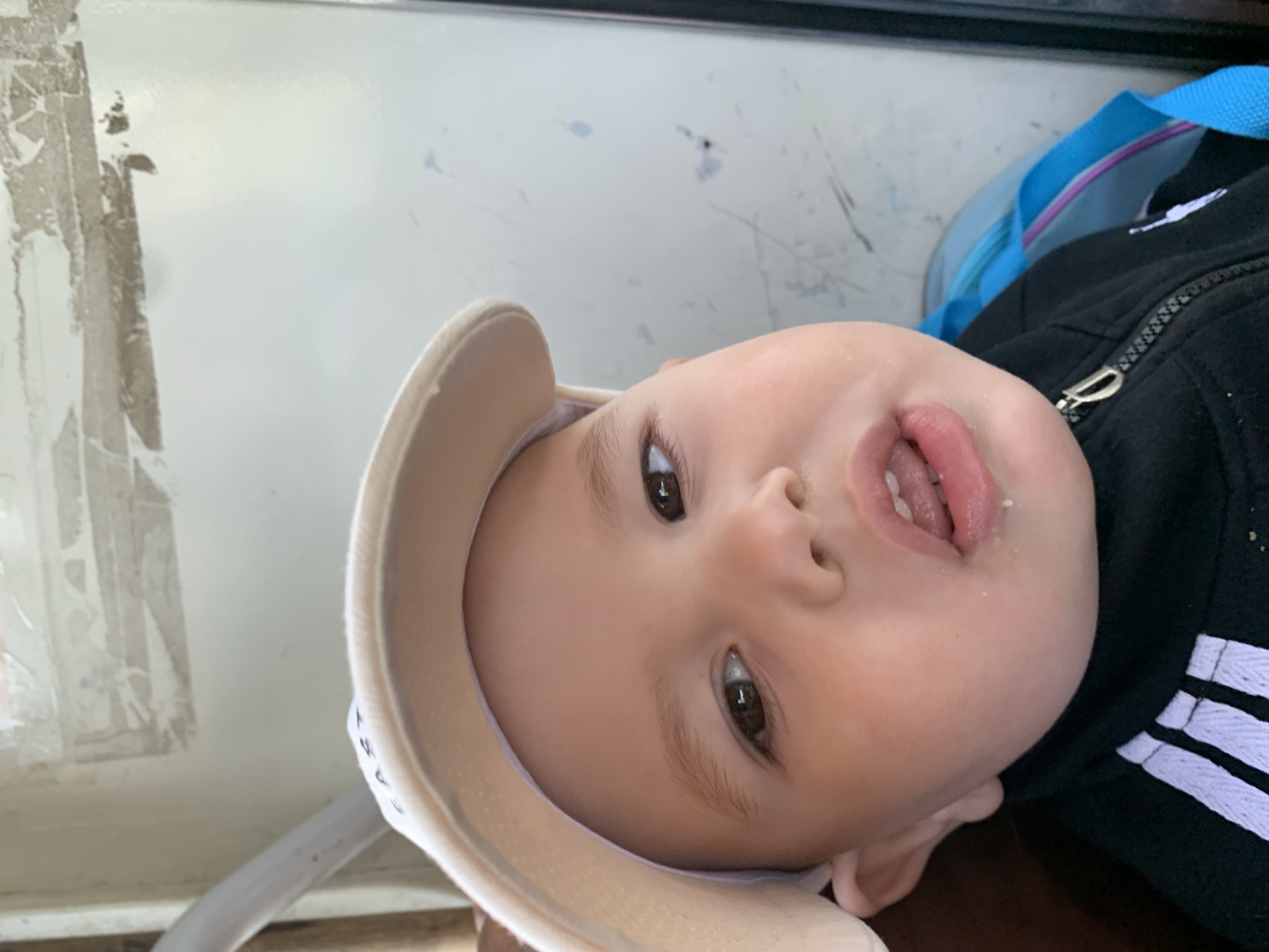 A close-up, tilted photograph features a baby wearing a light-colored sun visor and a black Adidas jacket with white stripes. The baby's mouth is slightly open, revealing the tongue and some small food particles. The background includes a white surface with scratches and a blue bag partially visible. The location is not discernible from the image. No specific details about the city of Rusalevka, Russia, are visible.