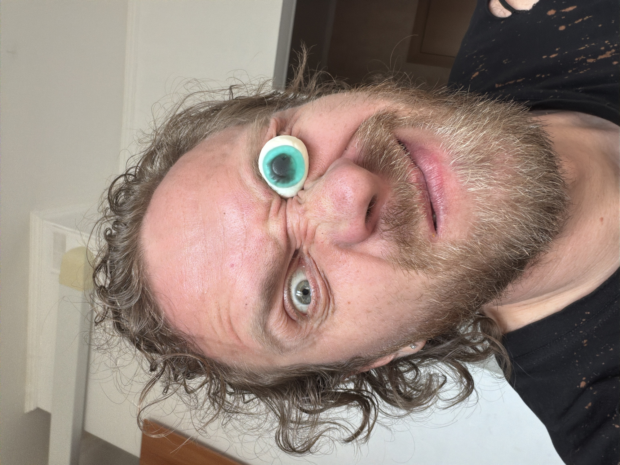 A Caucasian male with curly hair and a beard has a candy eyeball pressed against his forehead and nose. He is wearing a black t-shirt and is indoors. The location is unknown and there is no evidence that it is Saint Petersburg, Russia.