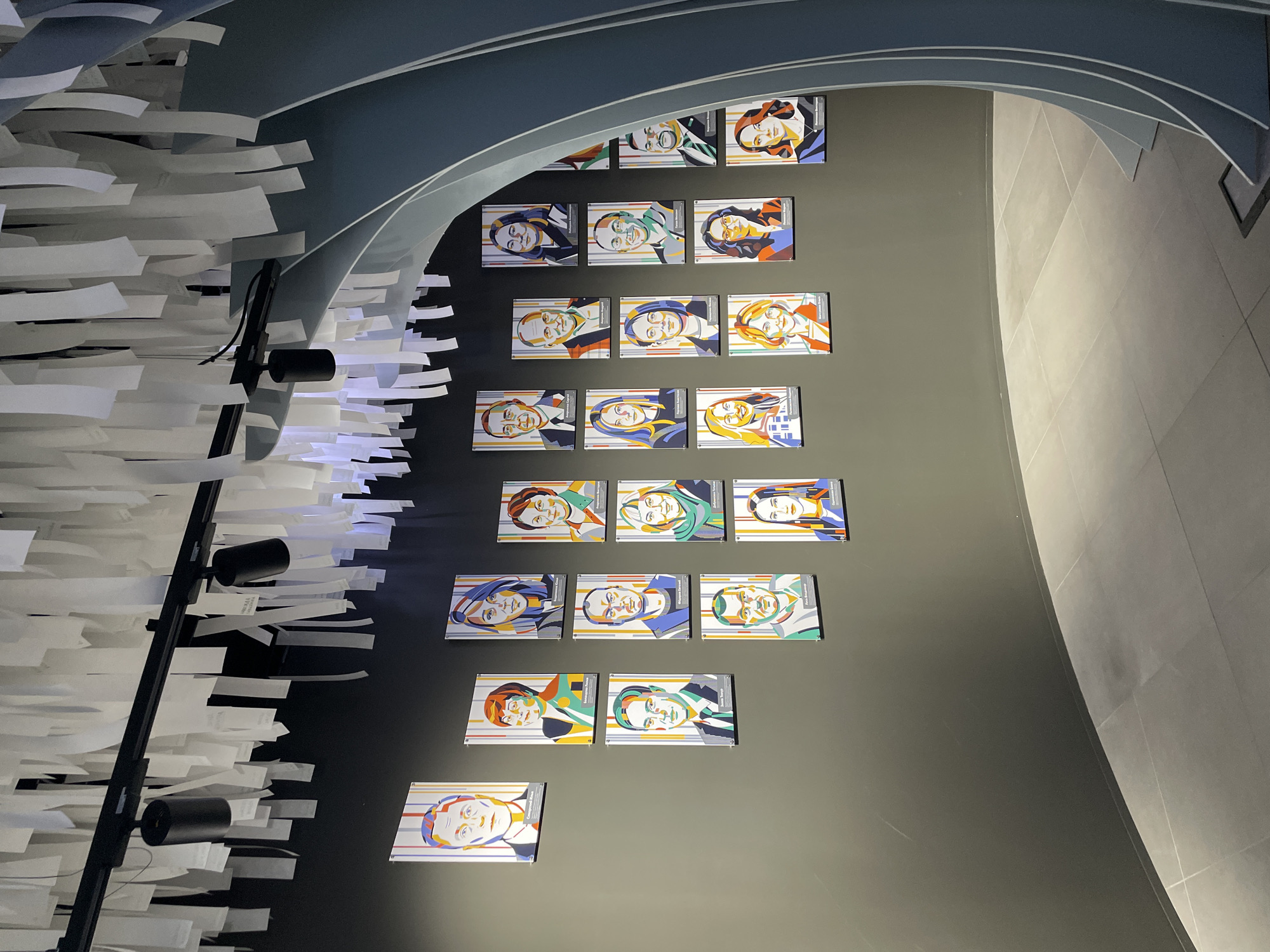 The image depicts an interior wall in Moscow, Russia, adorned with framed portraits arranged in a grid pattern. The portraits feature stylized depictions of individuals with distinct hair colors and clothing styles. The wall is painted in a neutral gray tone. A light fixture, consisting of a black metal frame and cylindrical lamps, is positioned on the left side of the wall. From the lamp protrudes white paper pieces. Overhead, a curved architectural element in shades of gray adds to the modern aesthetic of the space. The floor is covered with gray tiles.