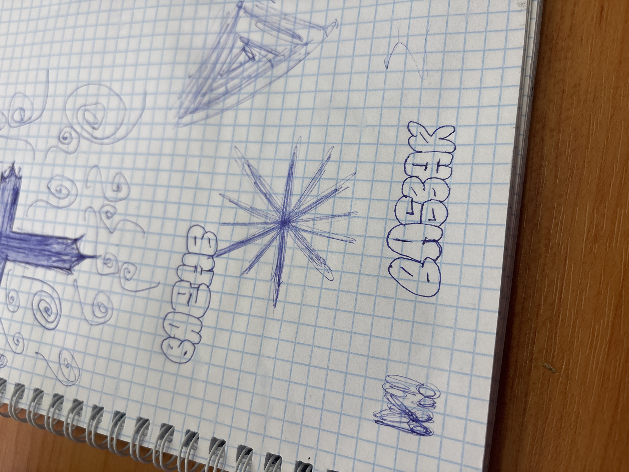 The image displays a close-up view of a spiral-bound notebook with grid paper, resting on a wooden surface. The notebook contains various doodles and drawings rendered in blue ink.
On the left side of the page, a prominent, dark blue, stylized cross or "X" shape is drawn, surrounded by a series of swirling and wave-like patterns. More faint, ethereal swirls extend upwards from this main drawing. In the central area, a distinct starburst or spiky circle design is visible, along with a fainter, abstract, geometric drawing above it that appears cone-like or triangular.
The word "СНЕЖОК" (SNEZHOK), which translates to "snowflake" or "snowball" in Russian, is written twice in a bubble-letter or graffiti style. One instance is on the left, partially obscured, and the other is more clearly visible towards the right side of the page. In the bottom right corner, a small, dense, abstract doodle of tangled lines is present.
The notebook's spiral binding is visible along the bottom edge, and the natural grain of the wooden surface underneath is apparent. The overall scene suggests a casual environment, likely a desk or table, where someone has been engaged in doodling or sketching. No individuals are present in the image.