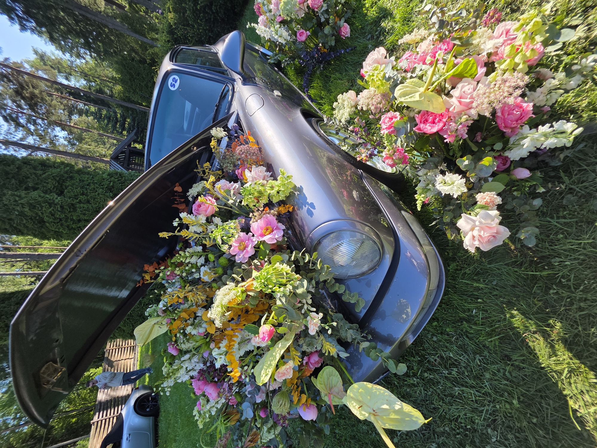 A gray car, possibly a Porsche, is parked on a grassy area in what appears to be Repino, Russia, indicated by a sticker visible on the car's windshield. The car's driver-side door is open, and the interior and exterior of the vehicle are filled with various types of flowers, including pink peonies, roses, and other colorful blossoms. Flowers also surround the front end of the car, partially obscuring the headlight. The background includes trees and a glimpse of a building or structure, suggesting an outdoor setting such as a park or garden.
