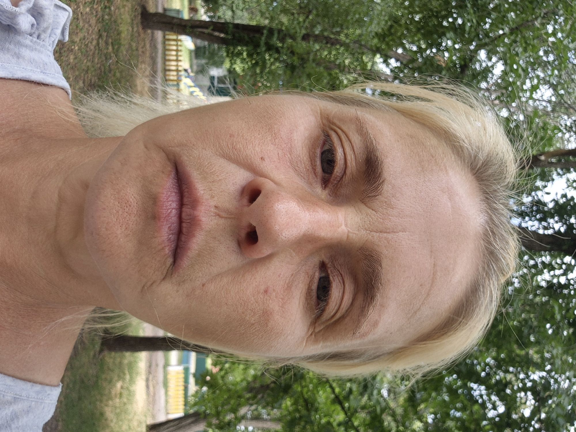 The image is a close-up, front-facing selfie of a woman with blonde hair, taken outdoors in Sozonivka, Ukraine. She has fair skin with visible wrinkles on her forehead, around her eyes, and on her neck. Her eyes are open and looking directly at the camera, with her gaze appearing somewhat tired. Her lips are closed, and her mouth is slightly downturned. The background consists of green trees with dappled sunlight filtering through, suggesting it is daytime. A wooden fence and a glimpse of a playground structure can be seen in the distance, indicating a park or recreational area. The woman is wearing a light gray t-shirt. The overall impression is a candid, unposed portrait capturing the woman in a natural outdoor setting.