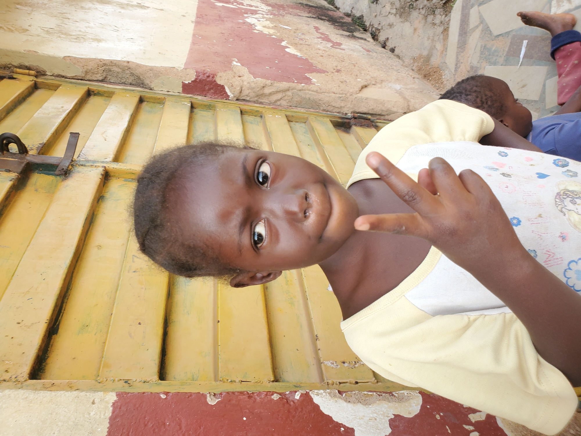 A young Black girl in a light yellow t-shirt is posed in front of a weathered yellow metal door and a textured, peeling red and tan wall. Her left arm is raised, and her index and middle fingers are extended in a peace sign. Her gaze is directed slightly to her left, away from the camera. Behind her, a younger Black child is seated, wearing a blue t-shirt and red pants, with their head bowed. The ground is paved with interlocking grey and tan tiles. The overall environment suggests an outdoor urban setting, likely in a residential or commercial area. The visual elements are consistent with a depiction of everyday life in the city of Jos, Nigeria.