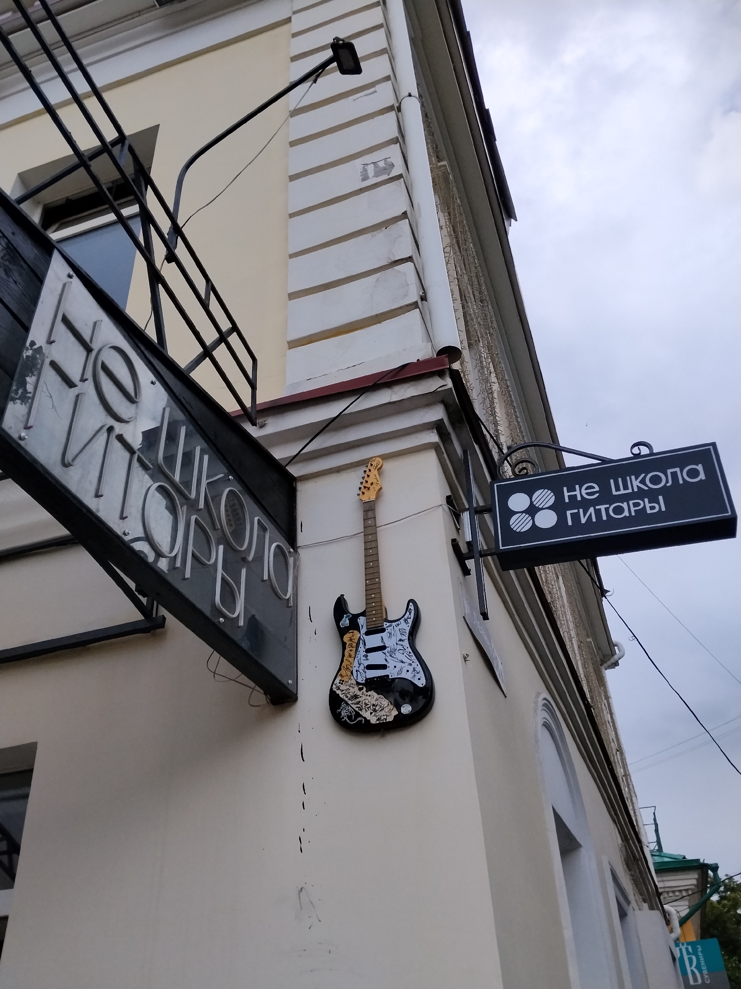 This media file captures an outdoor scene in Kaluga, Russia, featuring signage and a decorative element attached to the exterior of a building. The building appears to be old, with a light-colored facade and architectural details like pilasters.
The primary objects of interest are two signs and a decorative electric guitar. One sign, a large, dark rectangular sign with silver lettering, is mounted at an angle under an overhang. The text on this sign appears to be in Russian and reads "НЕ ШКОЛА ГИТАРЫ," which translates to "NOT GUITAR SCHOOL." To the right of this, a smaller, black rectangular sign with white lettering and a graphic emblem is also mounted on the building's corner. This sign also displays the text "НЕ ШКОЛА ГИТАРЫ."
A black electric guitar, adorned with various stickers and drawings, is mounted on the wall between the two signs. It serves as a whimsical decorative piece.
The scene is viewed from a low angle, looking upwards towards the building and the sky. The sky is overcast with clouds, suggesting a cloudy or possibly rainy day. The time of day is not clearly discernible, but the lighting conditions are consistent with daytime. The overall impression is a quirky and artistic facade for a place related to guitars, despite the sign humorously stating it is not a guitar school. There are no people visible in the frame. The setting is urban, indicated by the building and the presence of what appear to be utility lines or wiring connected to a light fixture.