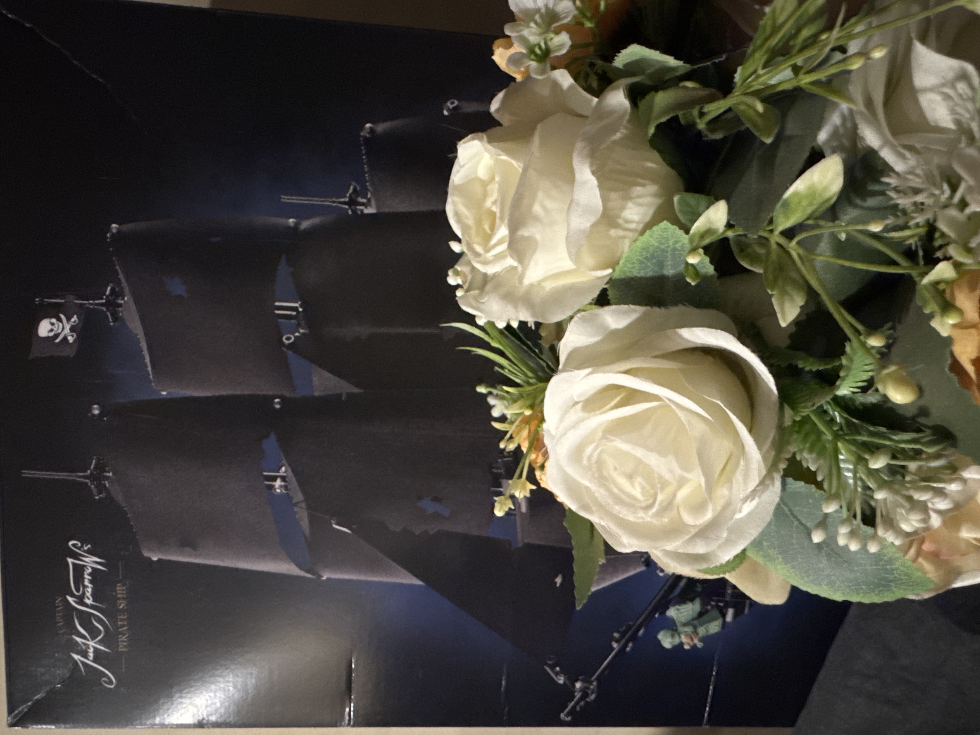The image depicts a close-up of artificial white roses and foliage in the foreground, with a dark background that appears to be a box or poster featuring a black pirate ship with a Jolly Roger flag. The ship is depicted sailing on dark water under a dark sky. Text on the box reads "CAPTAIN JACK SPARROW'S PIRATE SHIP." The lighting is dim, casting shadows and highlighting the textures of the roses and the glossy surface of the box. The scene is indoors, with no visible external environment or city details.