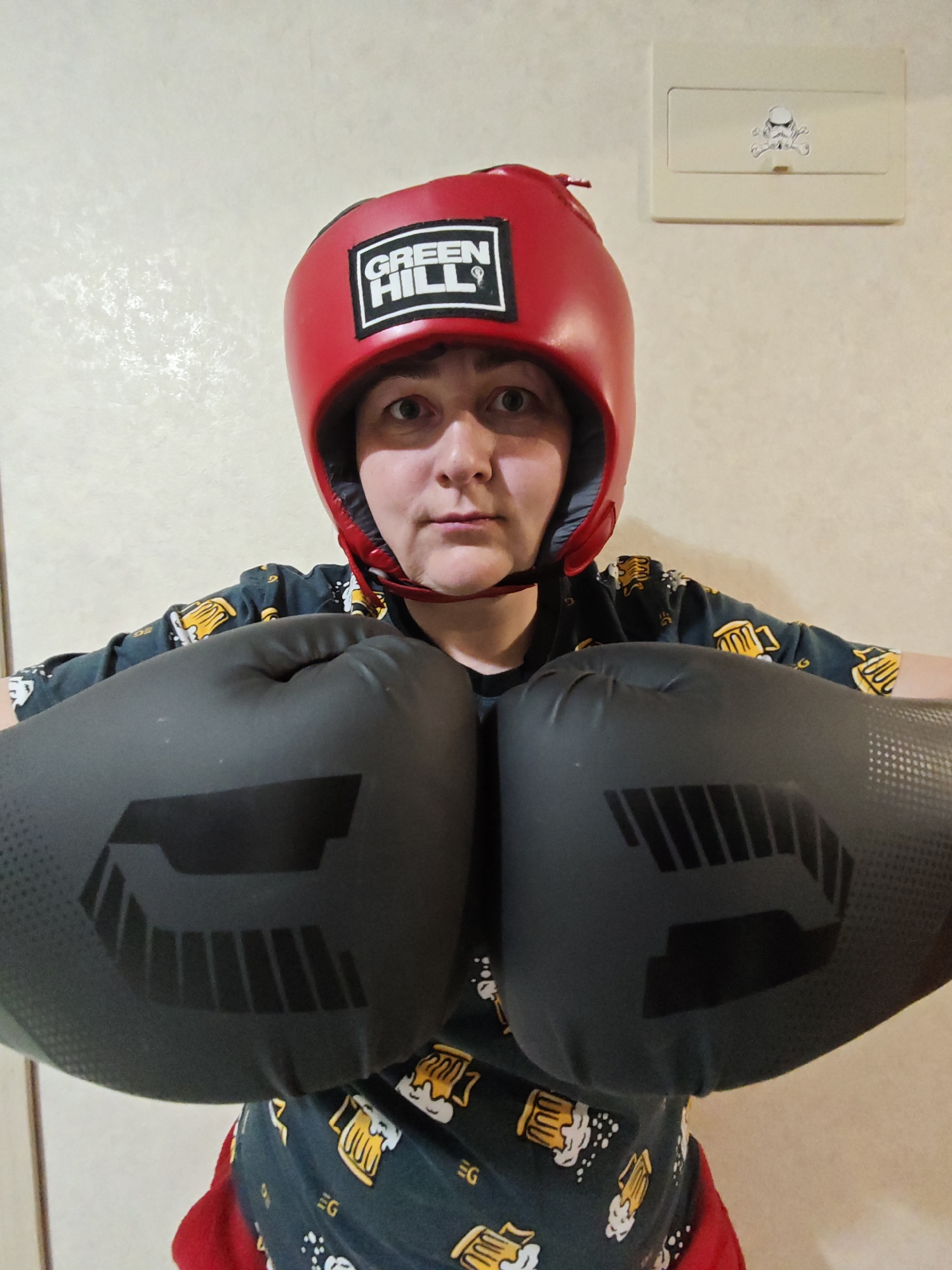 A person is centered in the image, wearing a red Green Hill boxing headgear and large black boxing gloves, positioned as if ready to box. They are wearing a dark green shirt with a pattern of beer mugs. The background is a plain, light-colored wall. A light switch with a sticker resembling a Star Wars Stormtrooper skull is visible on the upper right corner of the image. The photo was taken indoors, and the location is identified as Марковский, Russia.