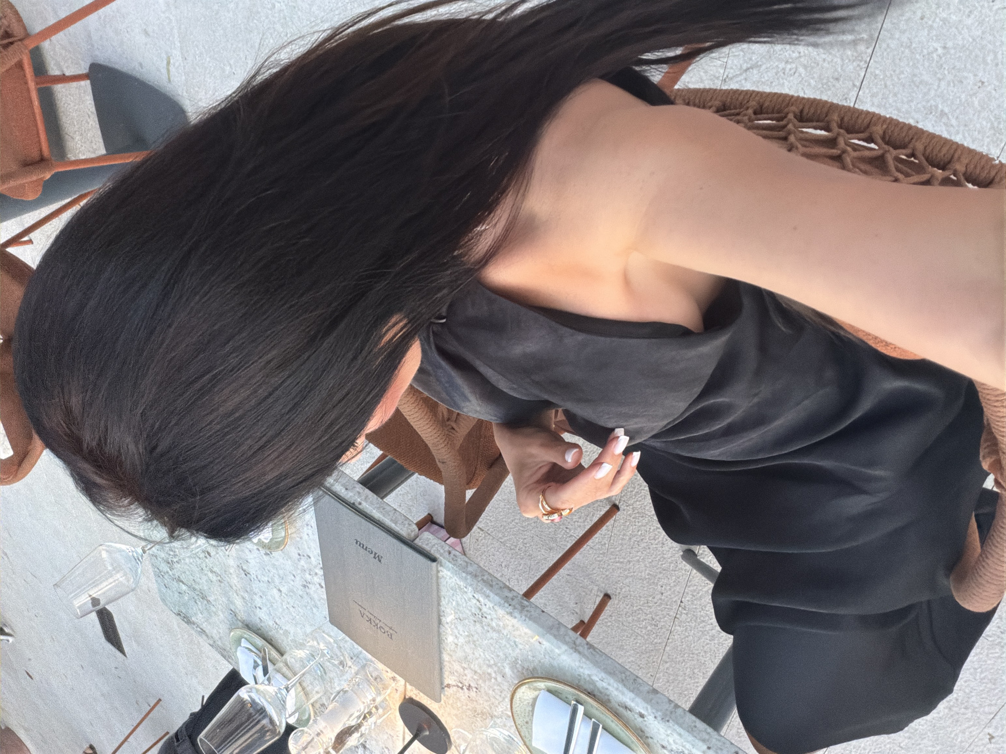 The image shows a person seated at a table with a menu. The person has long, dark hair and is wearing a black dress. They are looking down at the table, and their hands, adorned with rings, are visible. Several glasses are on the table, along with a menu that has "BOKKA" and "Menu" printed on it. The setting appears to be outdoors, with chairs and what looks like outdoor tiling visible in the background. The lighting suggests it could be daytime. The overall mood is relaxed and possibly indicative of dining or a casual meeting at a restaurant.