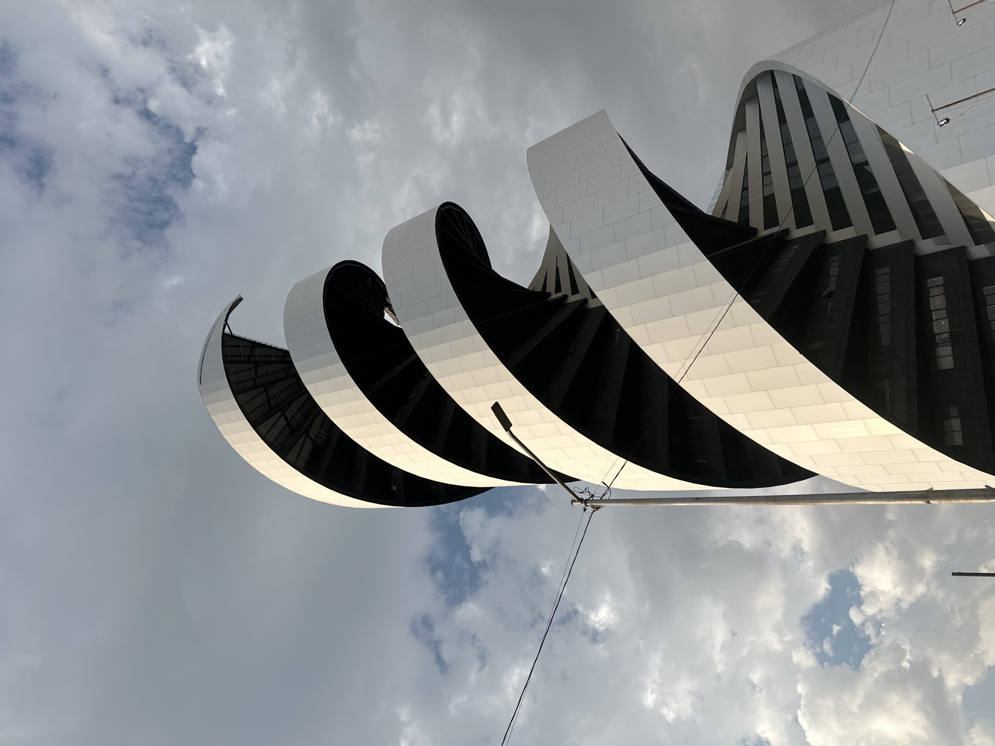 The image shows a low-angle view of the Museo Internacional Barroco (International Baroque Museum) in San Bernardino Tlaxcalancingo, Mexico. The modern architectural design features a series of white, curved structures that resemble ribbons or waves, contrasting with the dark, receding inner sections. The building partially faces a cloudy sky, which occupies a significant portion of the frame. A light pole and some wires are in the foreground, adding a sense of perspective to the architectural composition.