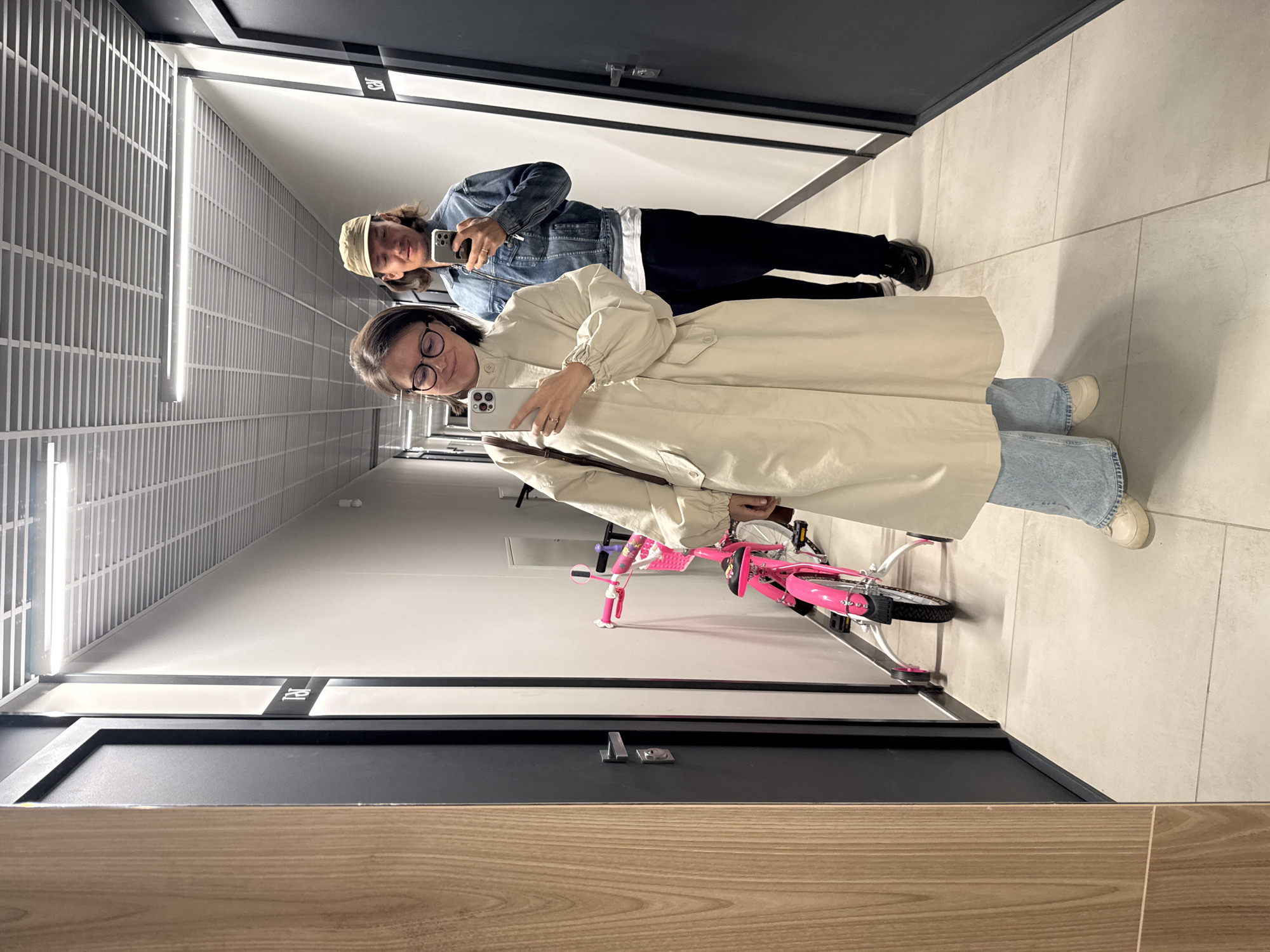 Two individuals are positioned in a brightly lit, modern hallway with doors lining either side. The hallway features a tiled floor and a suspended ceiling with linear lighting.
The person in the foreground is wearing glasses, a light-colored coat, and light-wash jeans, holding a smartphone. Behind them is a person wearing a denim jacket and a tan hat, also holding a smartphone.
A child's pink bicycle is leaning against the wall.
The location is Izhevsk, Russia.