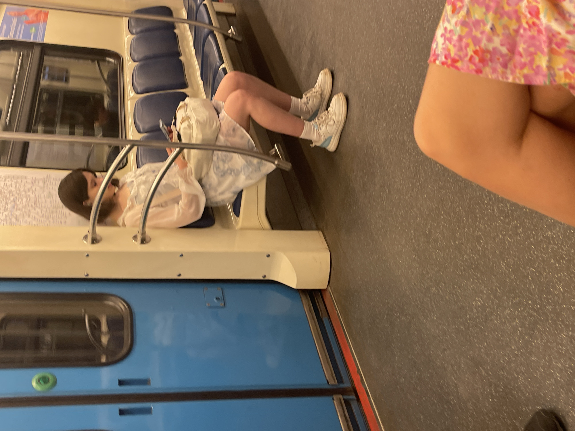 The image depicts the interior of a Moscow Metro train carriage. A young woman with brown hair is seated on a dark blue bench seat on the right side of the frame, looking down at a smartphone held in her hands. She is wearing a light-colored, possibly white or cream, long-sleeved top with ruffle or lace details, over a light-colored dress featuring a subtle blue and white pattern. A white face mask is visible, pulled down below her chin. A white shoulder bag rests on her lap. Her feet are clad in white ankle socks and white sneakers with light blue accents, resting on the dark grey, mottled floor.
The train interior features cream-colored wall panels, with horizontal silver handrails supported by cream-colored vertical stanchions. A row of dark blue, individual padded seats extends along the wall. Above the seats and handrails, a window reflects what appears to be another train interior or platform environment. A partially visible informational sign or map with Cyrillic script is mounted on the wall above the window.
On the left side of the frame, a bright blue sliding train door is visible, featuring a vertical window with rounded corners and a circular green button above two vertical recessed indentations. A thin red line marks the edge of the floor near the door. In the bottom right foreground, the bare legs and a floral-patterned skirt (pink, yellow, and white) of another individual are partially visible, indicating the photographer's perspective. A dark shoe or object is partially visible in the bottom left corner.
