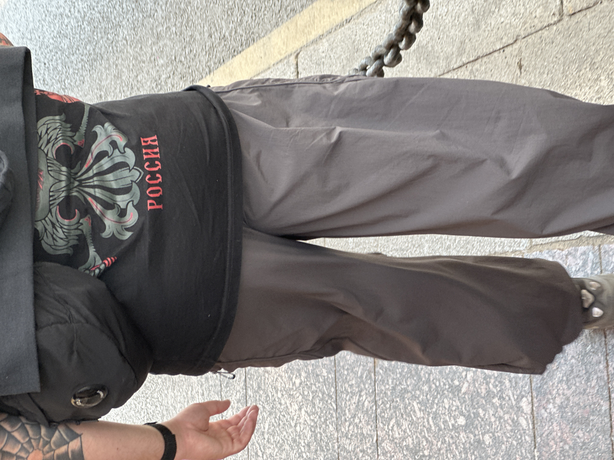 This image shows a person from the waist down, wearing dark pants and a black t-shirt with "РОССИЯ" (Russia) printed in red on it. The t-shirt also features a graphic design of what appears to be a stylized eagle or crest. The person is wearing a black jacket over the t-shirt and a black watch on their left wrist. A tattooed arm is visible in the lower left corner of the image. The background appears to be a textured stone surface with a yellow line and a thick metal chain, suggesting an outdoor urban environment. The lighting indicates it is daytime. The person is standing on a pavement or sidewalk. The overall impression is of a casual street scene in Saint Petersburg, Russia.