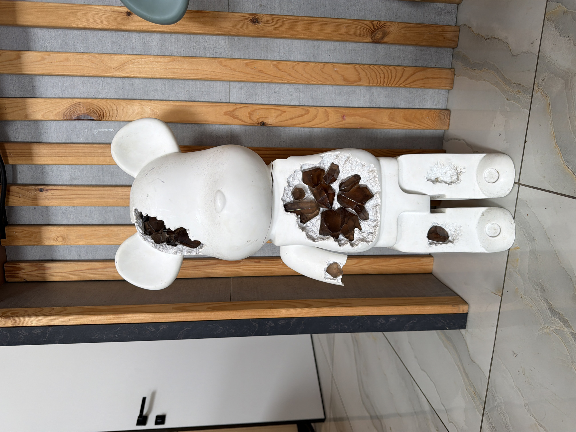 A white, bear-shaped figurine with broken sections revealing a brown, crystalline interior stands on a light-colored tiled floor in front of a wall adorned with vertical wooden slats. The figurine has damage to its head, chest, right arm, and legs. To the left of the figurine, a white door with black handles is visible. The environment appears to be indoors, possibly a residential or commercial space. The location is identified as Семилуки, Russia.