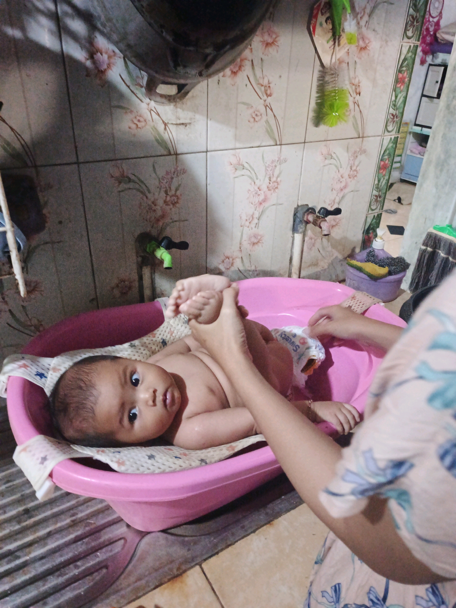 In Bumiharjo, Indonesia, a baby is being bathed in a pink plastic basin. The baby, wearing a diaper, is lying on its back on a white and patterned cloth within the basin. A person's hands are visible, holding the baby's feet. The basin is placed on a gray, ribbed surface, possibly a sink. The background features floral patterned tiles on the wall, with a faucet and a green bottle brush visible. The lighting suggests an indoor environment.