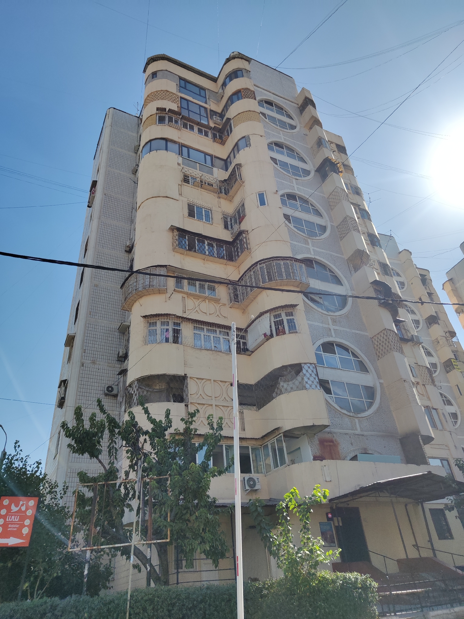 An upward-angle, medium shot captures a multi-story residential building with a unique, curved facade and large, circular windows. The building is painted in a pale yellow and grey color scheme, with decorative tile work in some areas. Several balconies with ornate metal railings extend from the building. The windows are mostly closed, and some have air conditioning units attached. A clear blue sky is visible above, with the sun shining brightly and creating lens flare. Overhead, a tangle of electrical wires crisscrosses the frame. In the foreground, lush green trees and bushes partially obscure the lower portion of the building. A red and white sign with some text and an arrow is visible on the left side of the frame. The building is located in the city of Tashkent, Uzbekistan.