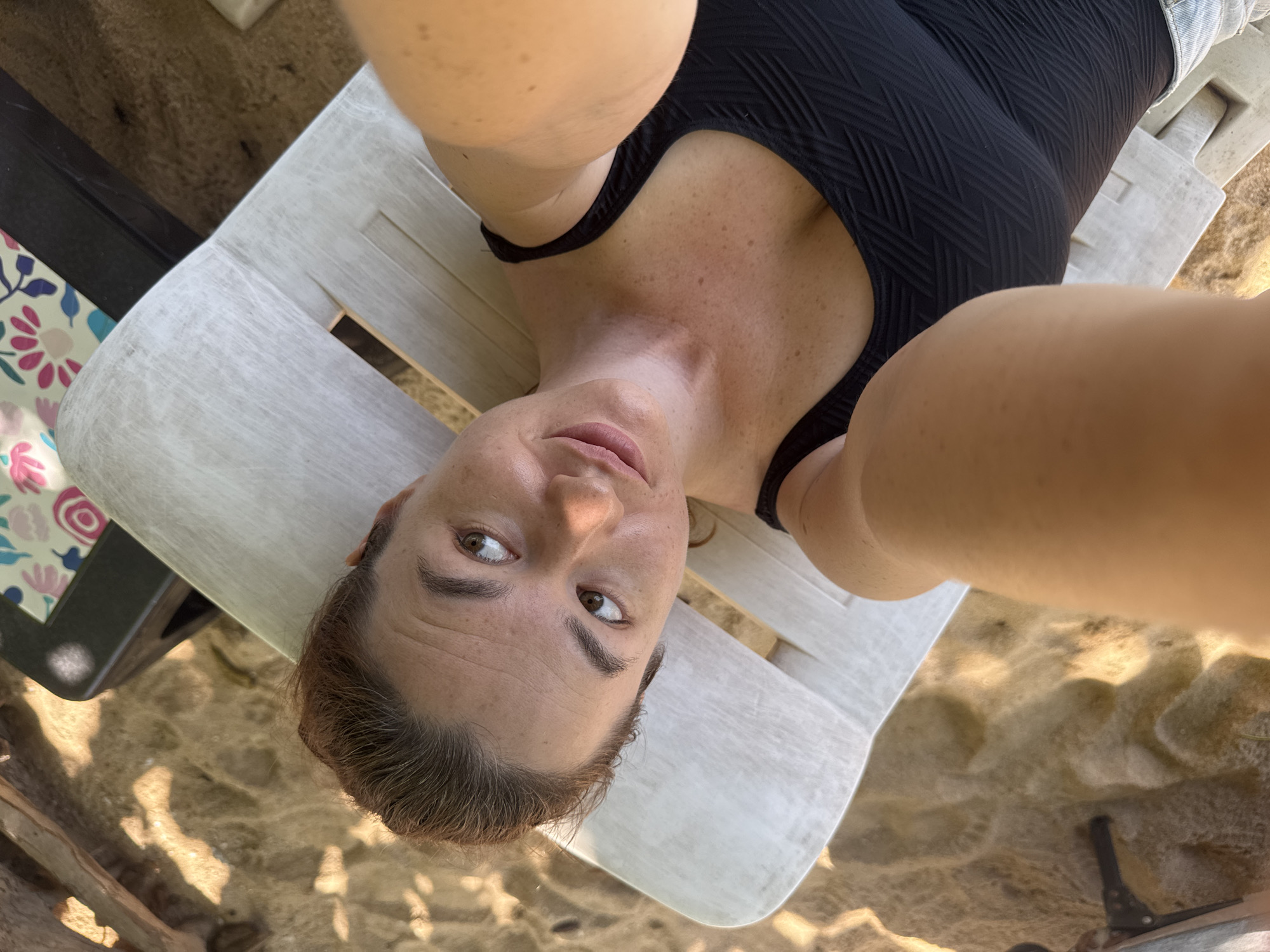 A high-angle, close-up, selfie-style shot captures a woman lounging on a white plastic beach chair in Rawai, Thailand. The woman, with light brown hair pulled back, is looking up and to her left, her face partially illuminated by sunlight. She wears a black one-piece swimsuit with a textured pattern. Her bare shoulders and décolletage are visible, sprinkled with freckles. The beach chair is situated on a sandy surface, with scattered debris and the edge of what appears to be a patterned cooler visible in the background. Sunlight casts dappled shadows on the sand.