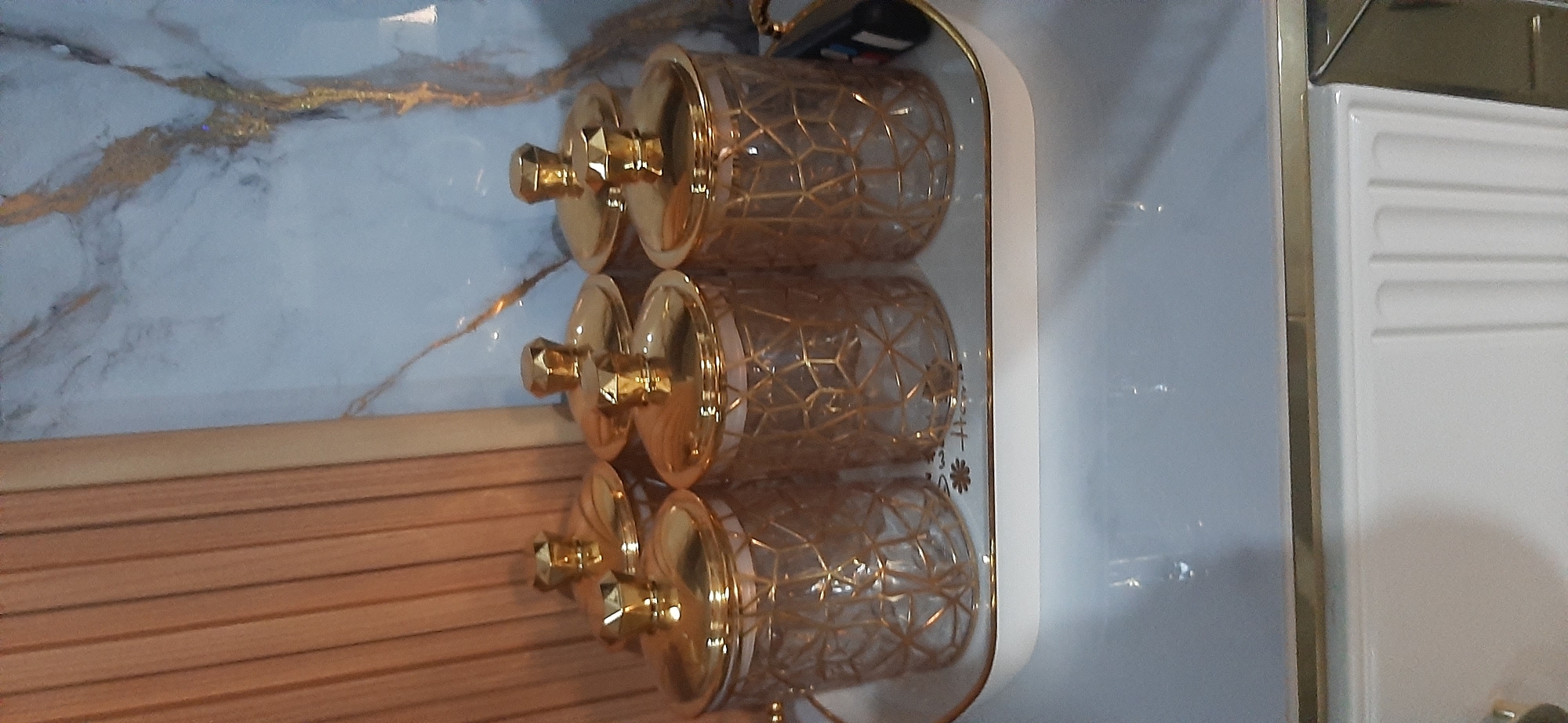 A set of six glass jars with gold geometric patterns and gold lids are arranged on a white tray with gold trim. The tray is placed on a white surface. In the background, there is a wooden paneled wall and a marble-patterned wall with gold veins. The image is taken from a slightly high angle. The specific location in Jambi, Indonesia is not determinable from the visual information provided.