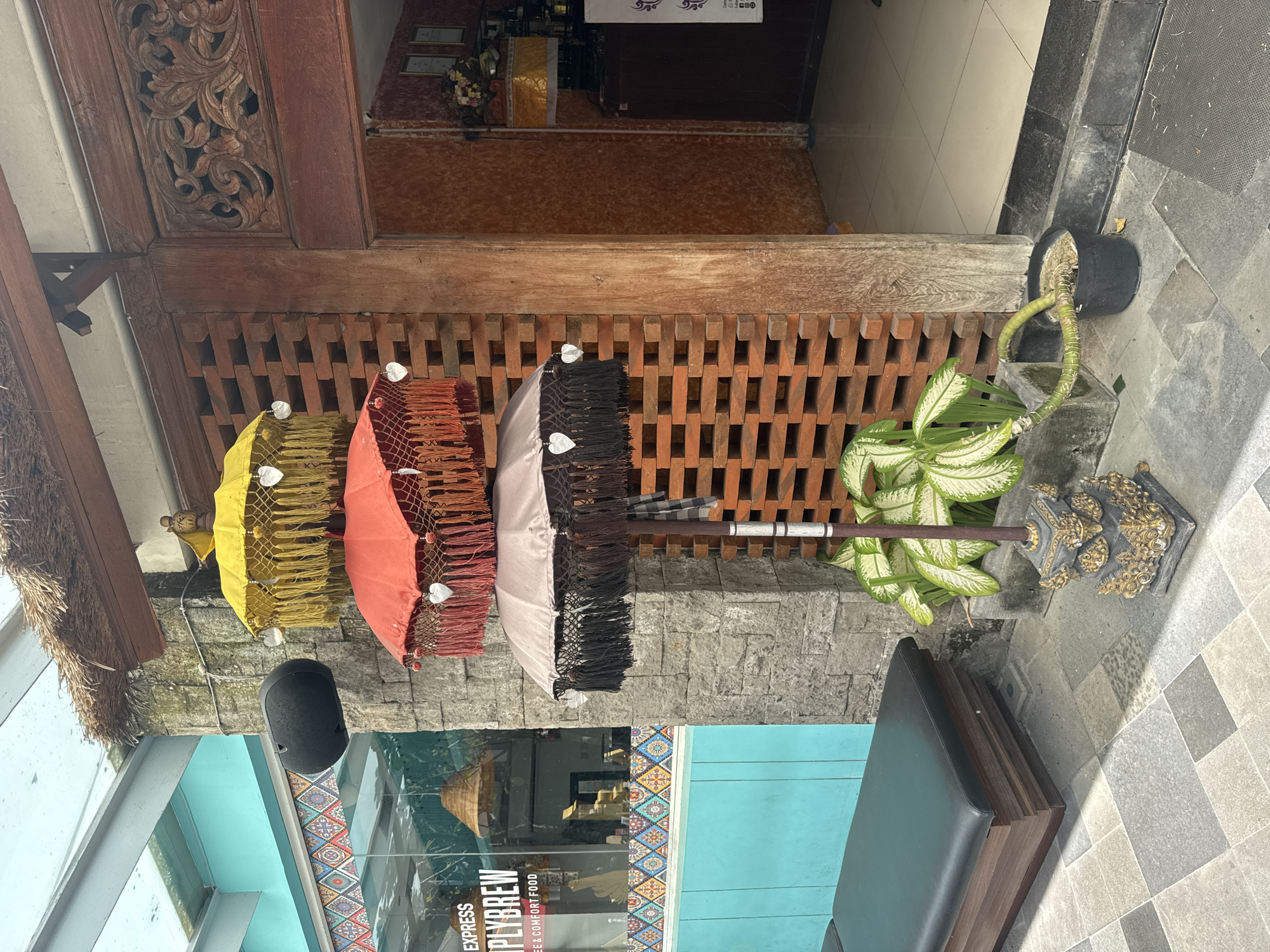 The image depicts a street scene in Kuta Selatan, Indonesia. Four decorative umbrellas – yellow, coral, light gray, and black – hang from a wooden structure above a storefront. The store's glass facade reflects the interior, visible through the lettering "EXPRESS PLYBREW COFFEE & COMFORT FOOD". Adjacent to the wooden structure, a potted plant stands next to a bench, and a speaker is mounted nearby. The surrounding area features tiled walkways.