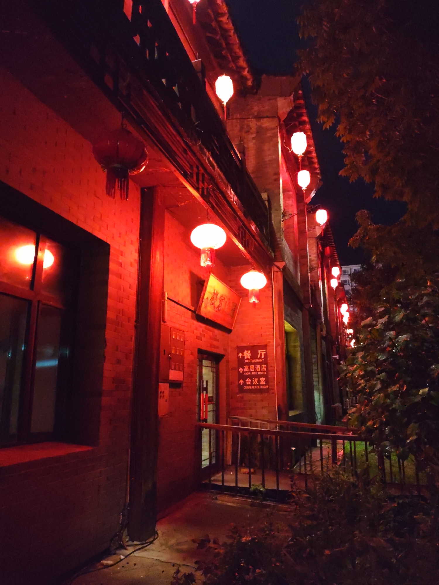This is a nighttime, low-angle shot of a brick building illuminated by red lanterns and other red lighting. The building appears to be a commercial establishment, with a sign indicating "RESTAURANT," "HIGH-RISE HOTEL," and "CONFERENCE ROOM." Several white, spherical lanterns hang from the building's facade, casting a bright glow. Red Chinese lanterns are also visible. The building has traditional Chinese architectural elements, including a dark wooden balcony or eaves. A dark-colored railing and a set of stairs lead up to an entrance. Lush green foliage is present to the right of the building, partially obscuring a portion of the structure and its lanterns. The overall impression is one of a vibrant and inviting scene, possibly in a traditional district of 林甸镇, China.