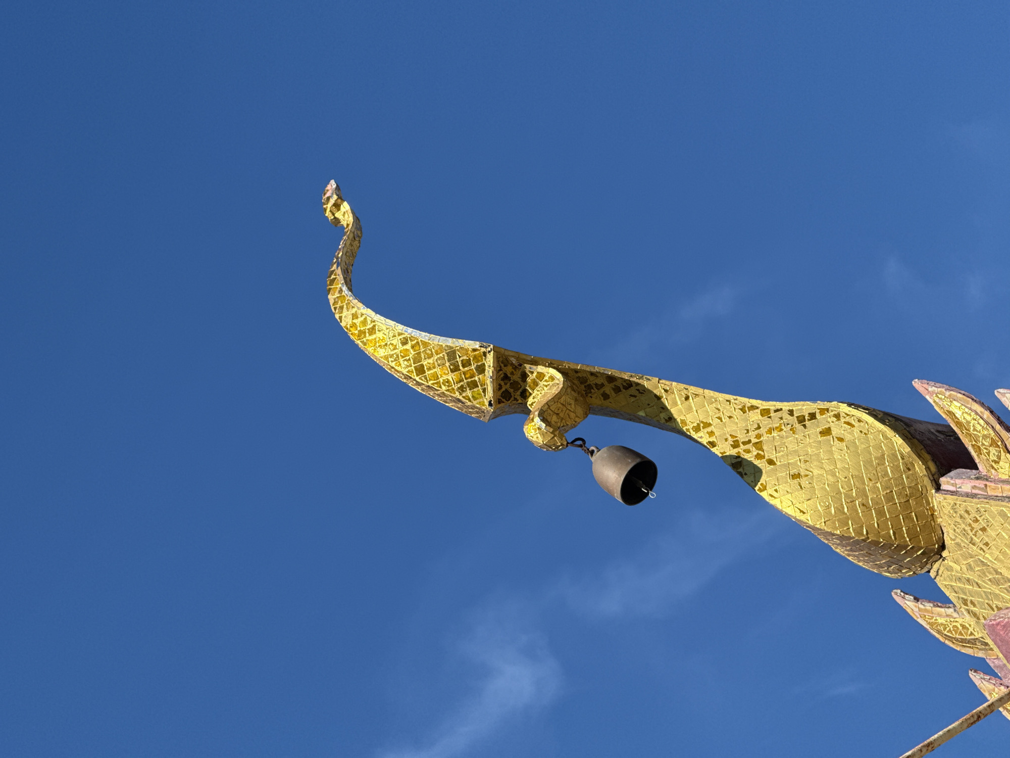 This is a close-up, low-angle outdoor shot of the gilded roof finial of a temple building in Baan Plai Laem, Thailand. The ornate finial, which appears to be stylized in the shape of a mythical creature, is covered in small, shimmering golden mosaic tiles. A small, dark-colored bell hangs from a chain attached to the finial. The background is a clear, vibrant blue sky with a few wispy white clouds. The focus is sharp on the finial and the bell, with the sky slightly less defined. No people are visible in the frame.