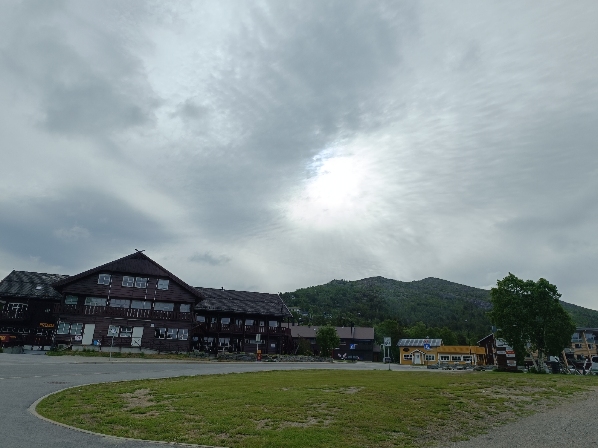 The image depicts a view of a village in Hovden, Norway, under a cloudy sky with patches of sunlight breaking through. In the foreground, there is a grassy area and a paved road. A large, dark wooden building with multiple windows and balconies dominates the left side of the image. Signs indicating "PIZZABAR" are visible on this building. To the right, there are smaller, colorful buildings, including a yellow one with a white frame and a dark brown building with signs. Behind the buildings, a densely forested mountain rises, with some houses scattered on its slopes. A tall green tree stands in the middle ground on the right. The overall scene suggests a commercial or residential area in a mountainous, potentially tourist-oriented location.