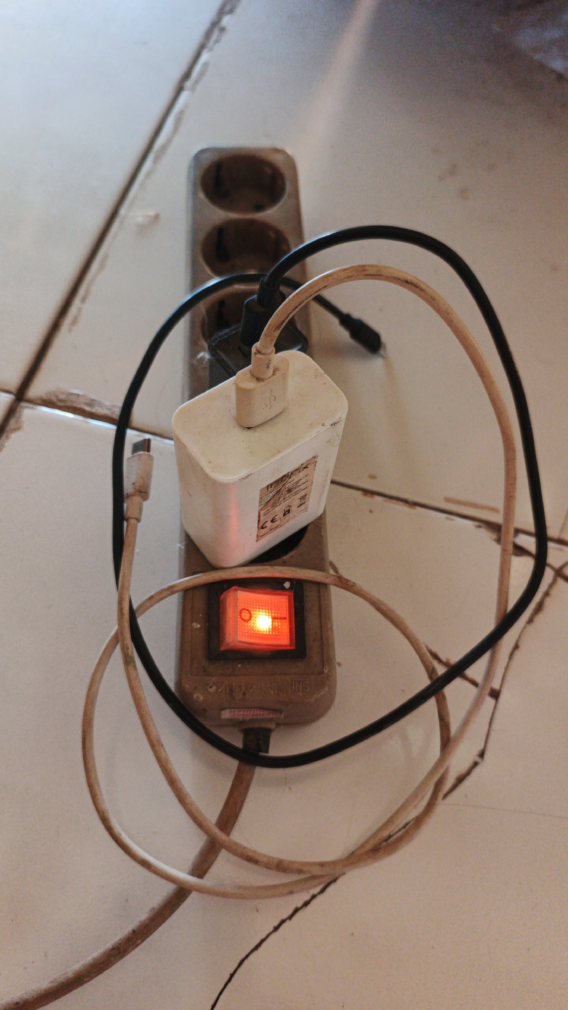 An overhead, close-up shot shows a power strip with two outlets, plugged into an unseen electrical source on a light-colored tiled floor. A white AC adapter is plugged into the upper outlet and is connected to a beige USB cable. A small, red illuminated switch is present on the power strip. A black wire and another beige wire are loosely coiled around the power strip and adapter. The scene is consistent with a residential or office setting in Santing, Indonesia, where such electrical accessories are common for charging devices.