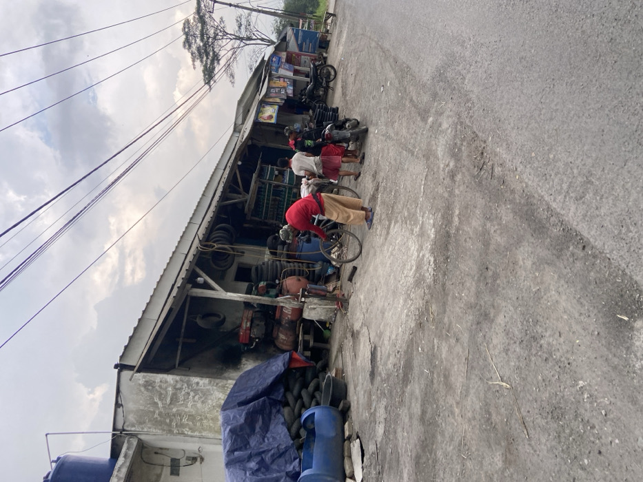 The image depicts a street scene in Kecamatan Margaasih, Indonesia, a small town located in the Bandung Regency. The scene is captured from a perspective that looks down the street, with the camera positioned in front of a roadside shop. The shop, which appears to be a motorcycle repair or tire shop, has a weathered concrete facade and an open front. Stacks of tires can be seen inside the shop, and there are also a few items of machinery, including an air compressor and a workbench. Several people are gathered around the shop. Two men stand in front of the shop, one in a red shirt and the other in a blue shirt. They appear to be talking. A third man, also in a red shirt, is crouched down beside a bicycle with its back wheel removed. He is working on the bike. Two other men stand behind the first two men, watching the man with the bike. They are wearing casual clothing. The street is made of asphalt, and it is largely empty. Several power lines run overhead, and there is a small tree in the distance. The sky is overcast, and the overall atmosphere is subdued.