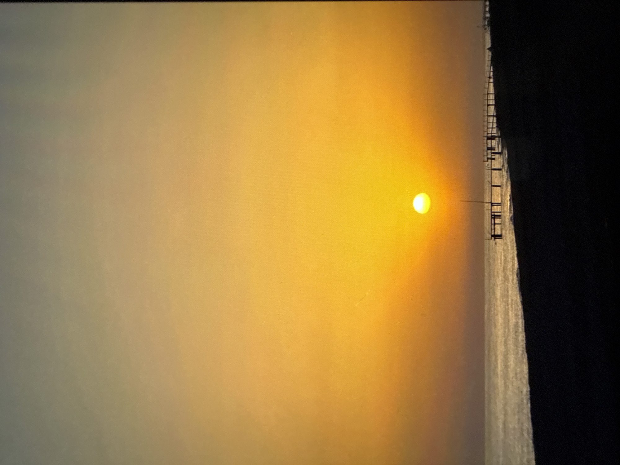 The image depicts a beach scene at sunset. The sun, a bright orb, is positioned low in the sky, casting a warm orange and yellow glow across the horizon. A long pier extends from the shore into the calm sea, with several silhouetted figures walking along it. The foreground is a dark, sandy beach. The scene is devoid of any specific landmarks that would indicate a precise location within Mykolaiv, Ukraine.