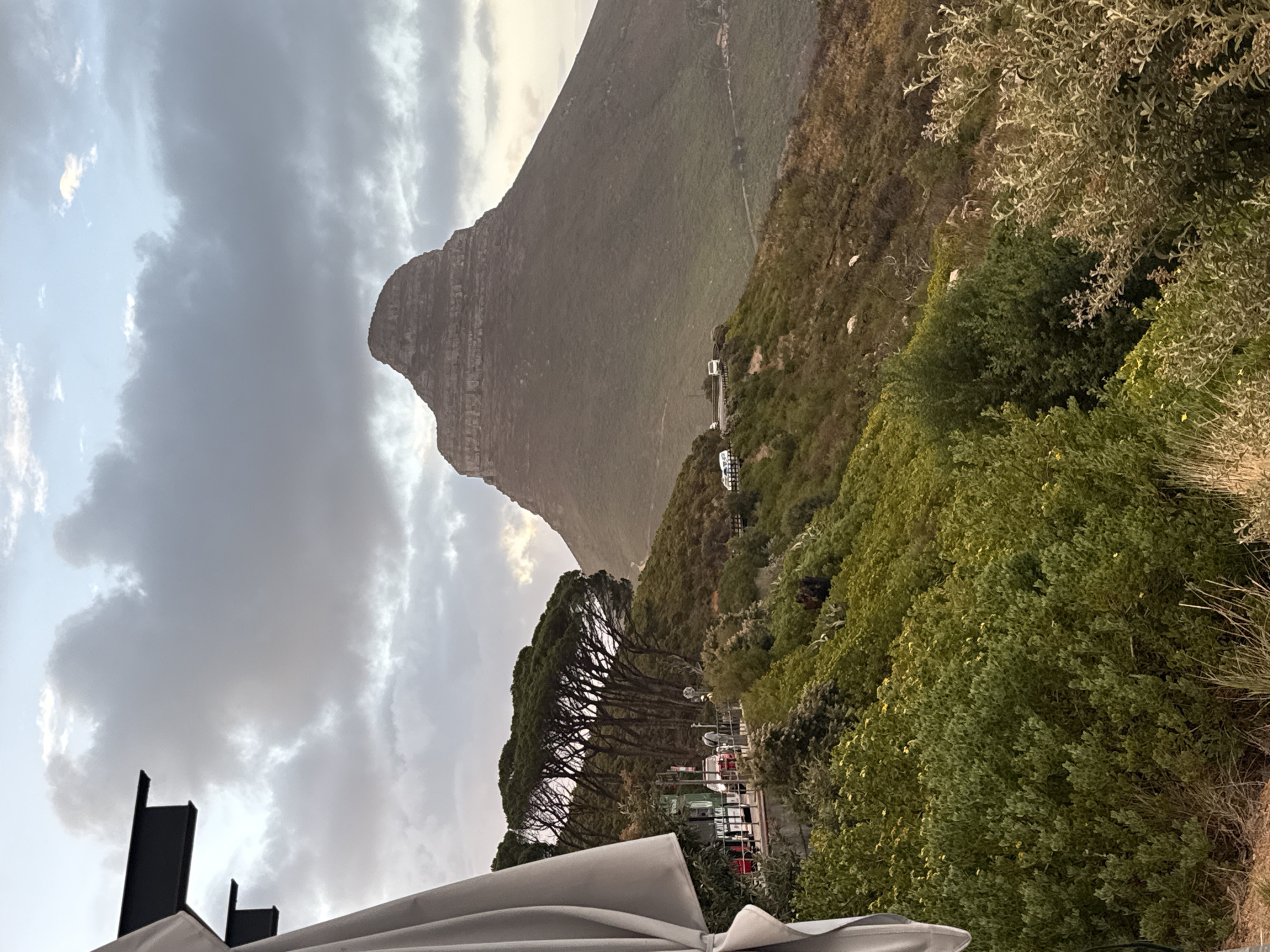 The image shows a view of Lion's Head mountain in Cape Town, South Africa, under a cloudy sky. The mountain is in the background, with distinct layered rock formations visible. A road with parked vehicles winds up the side of the mountain. In the foreground, there's dense vegetation including trees and bushes. A structure, possibly part of a building, with a white awning is partially visible at the bottom of the frame.