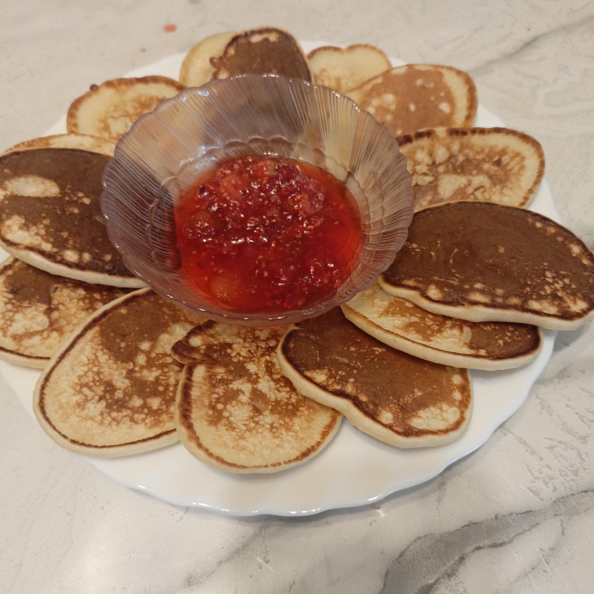 A top-down shot showcases a white plate laden with numerous small, round pancakes, arranged in a circular pattern around a clear, fluted glass bowl. The pancakes exhibit varying degrees of golden-brown to darker brown coloration, indicating a cooked surface. The central glass bowl is filled with a vibrant red, chunky compote or jam, likely raspberry or strawberry, with visible fruit pieces. The white plate, which has a subtly scalloped or wavy edge, rests on a light-colored surface, possibly a countertop, with subtle grey marbling or pattern. The scene depicts a prepared meal or snack, likely breakfast or dessert, presented for consumption. No people or visible text are present. The lighting is soft and even, suggesting an indoor setting.