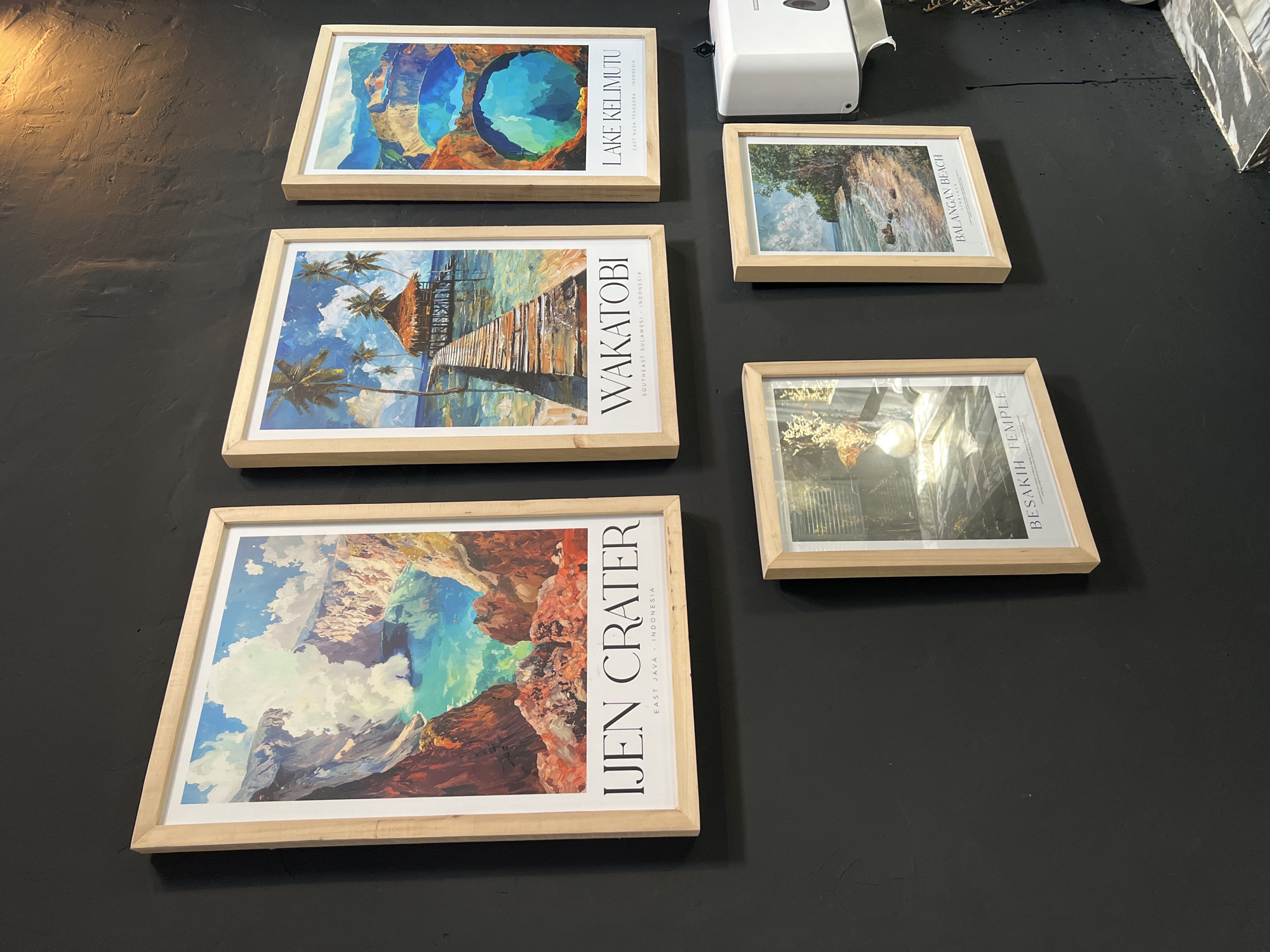 The image shows five framed pictures lying on a dark surface. From left to right, the pictures depict "IJEN CRATER" (East Java, Indonesia), "WAKATOBI" (Southeast Sulawesi, Indonesia), "LAKE KELIMUTU" (East Nusa Tenggara, Indonesia), "BALANGAN BEACH", and "BESAKIH TEMPLE". A white hand sanitizer dispenser is visible in the upper right corner, along with some decorative items. The scene appears to be set in a location in the city of North Kuta, Indonesia, as suggested by the images of Indonesian landmarks.
