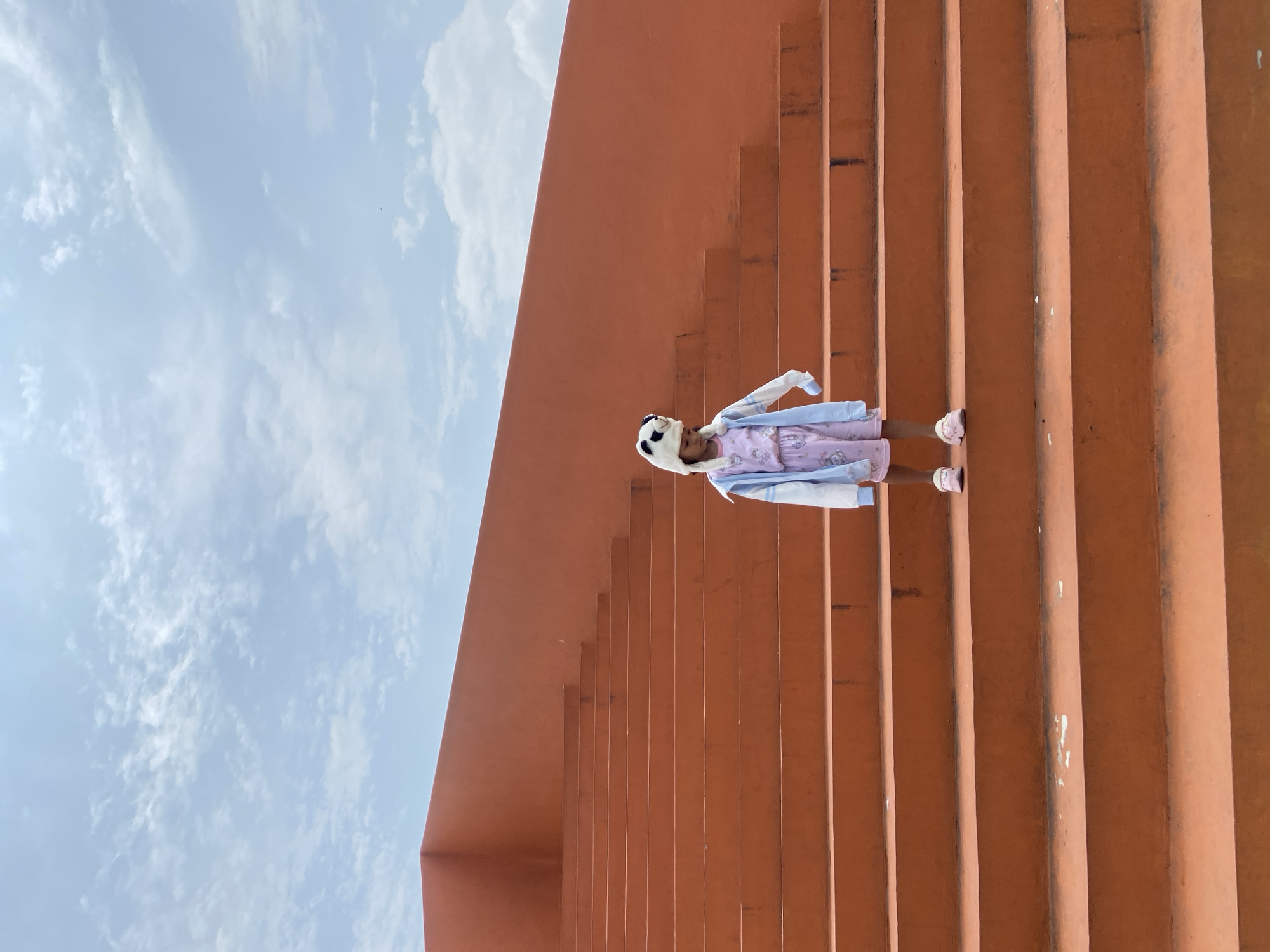 The scene depicts a young child standing centrally on a wide outdoor staircase within the city of Sidoarjo, Indonesia. The child, wearing a pink patterned pajama set, a light blue and white long-sleeved jacket, and a white panda-themed hat, is positioned on the fifth step from the bottom, looking downwards. Her feet are shod in pink slip-on shoes. The staircase and an adjacent angular wall are constructed from concrete and uniformly painted in a matte terracotta or orange-brown color. The stairs ascend from the lower left to the upper right of the frame, showing minor scuff marks. The upper portion of the image reveals a light blue sky with scattered white clouds, indicating a daytime setting. No other individuals or distinct objects are present.