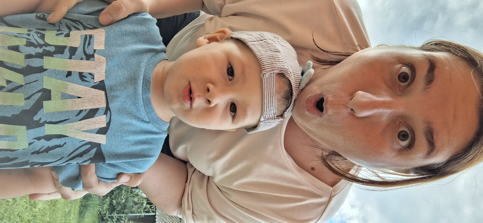 A close-up, low-angle selfie shows a woman and a young boy outside. The woman, with brown hair and wide eyes and an open mouth, leans in from behind the boy. She wears a light-colored t-shirt. The boy, wearing a striped baseball cap and a blue t-shirt with the text "STAY WILD" and a tiger graphic, looks directly at the camera. His hands are resting on his chest. The background shows a cloudy sky and some greenery, suggesting an outdoor setting, potentially a residential yard in Chaikovsky, Russia, based on the provided context. The overall atmosphere appears playful and informal.