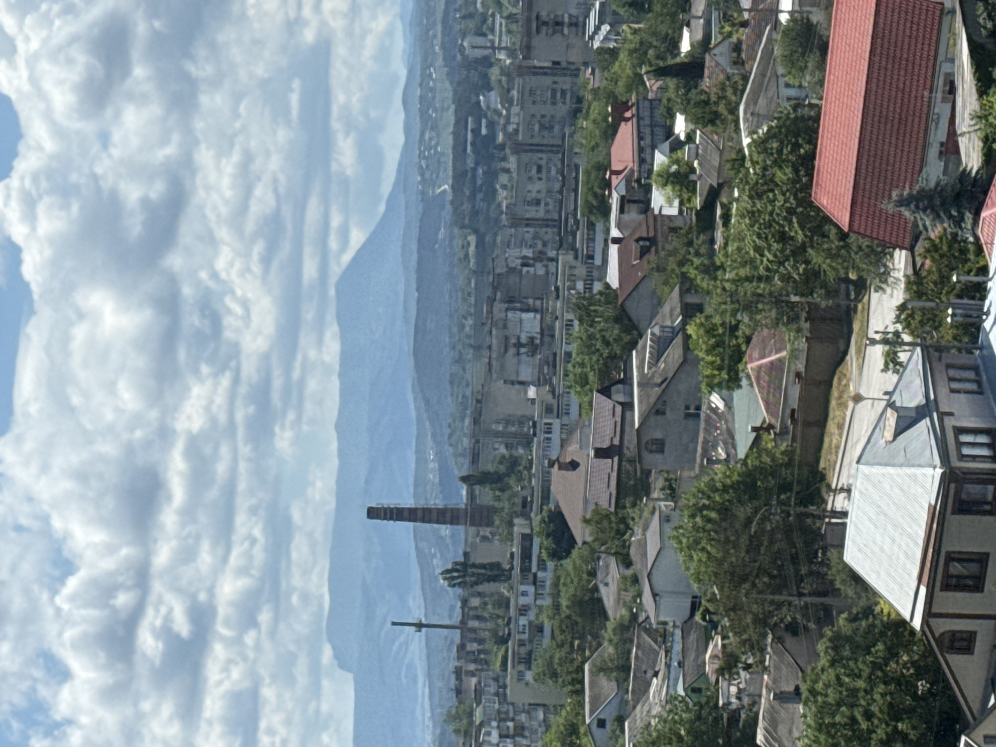 The image captures an elevated, panoramic view of a town under a cloudy sky. The foreground is dominated by the roofs of residential houses, varying in color and material, with some featuring prominent chimneys. Dense greenery, including trees and shrubs, is interspersed throughout the residential areas.
Further into the scene, apartment buildings and other structures are visible, suggesting a more urbanized section of the town. A tall, brick chimney stands out as a prominent industrial element amidst the buildings. In the background, a range of mountains recedes into the distance, partially obscured by the atmospheric haze and clouds. The overall impression is one of a moderately sized town with a mix of residential and industrial areas, nestled within a landscape of rolling hills and mountains. No people or specific actions are discernible in the image.