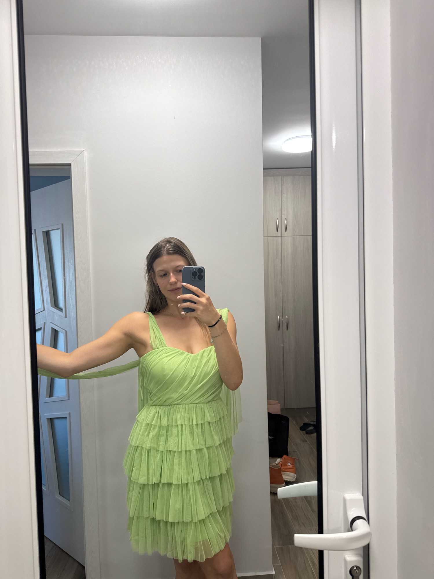 A woman is taking a mirror selfie in a room with a white door and wooden cabinets. She is wearing a lime green ruffled dress and holding a cell phone. In the reflection, a light fixture is visible on the ceiling, as well as some shoes and bags on the floor. The room appears to be a residential interior. The location is not specified to be in Sofia, Bulgaria, but rather a general interior residential setting.