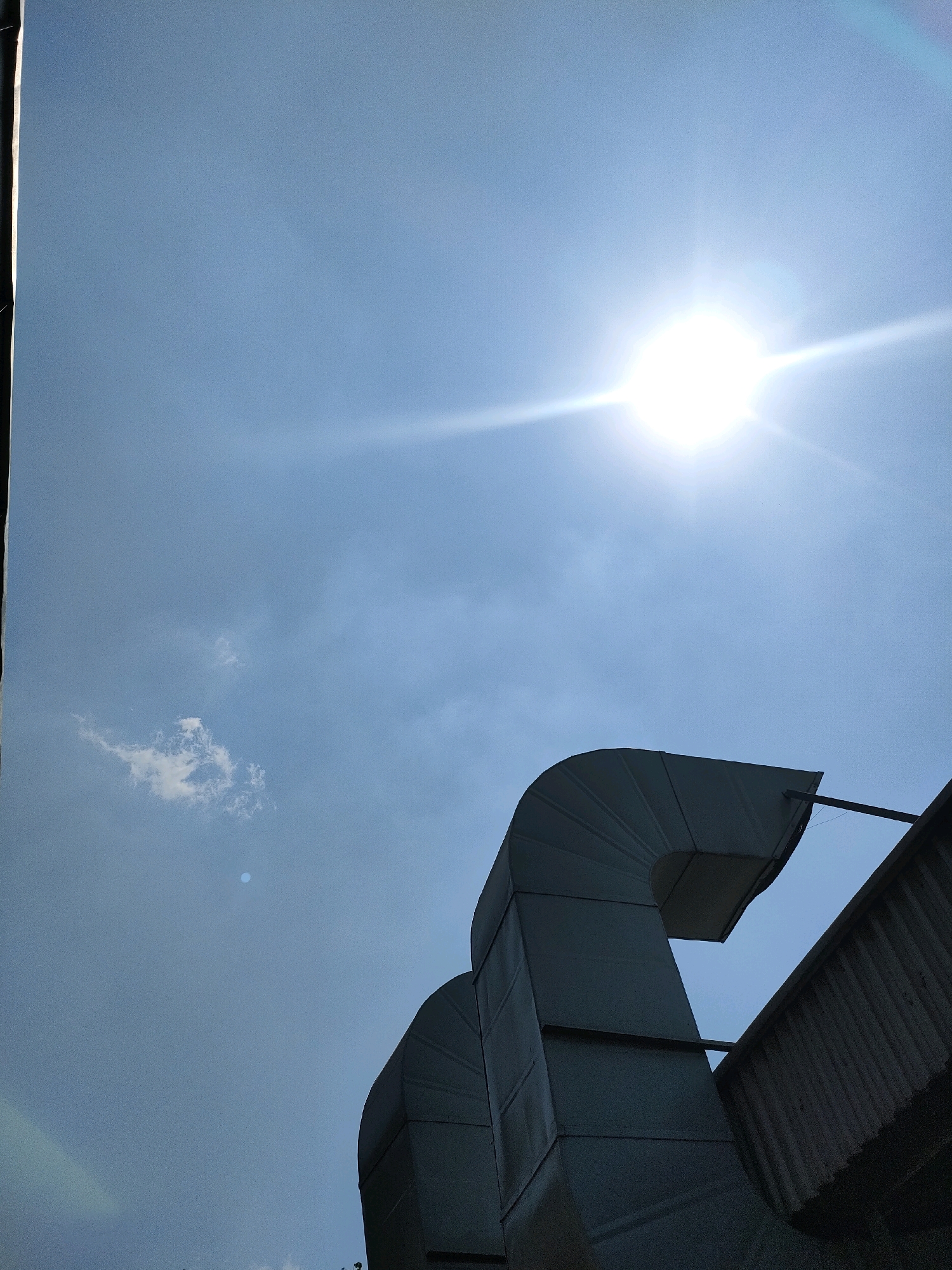 The image shows a bright sun shining in a clear blue sky, with a few wispy clouds. In the foreground, there are two large, gray ventilation ducts made of metal, positioned at an angle. A corrugated metal roof is visible to the right of the ducts. The scene appears to be outdoors, likely on a rooftop, possibly in an industrial or commercial area. There are no people or animals visible in the image. The sunlight is intense, creating a lens flare effect in the sky. The overall impression is one of a bright, sunny day, with industrial infrastructure dominating the lower part of the frame. There is no specific indication of the location within Sayung, Indonesia, but the presence of industrial elements suggests an area that may be involved in manufacturing or commerce.
