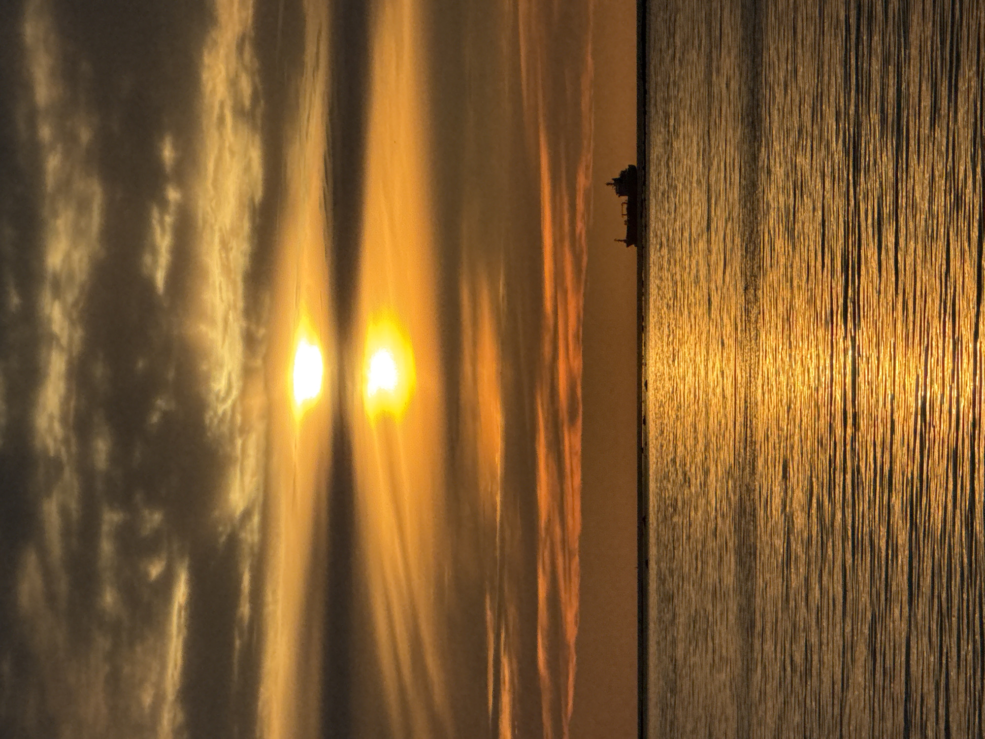 The image depicts a serene seascape at dusk, with the sun low on the horizon casting a warm, golden glow. The sky is a tapestry of layered clouds, ranging in color from deep gray to bright orange, with the sun appearing as a brilliant, diffused orb. The sea stretches out in the foreground, its surface rippled by gentle waves, reflecting the fiery hues of the sky. A lone cargo ship is visible in the distance, silhouetted against the bright horizon, adding a sense of scale and tranquility to the scene. The overall impression is one of peace and natural beauty. The location is the Baltic Sea, likely viewed from the coast of Tallinn, Estonia, possibly near the Old City harbor or a more remote beach.