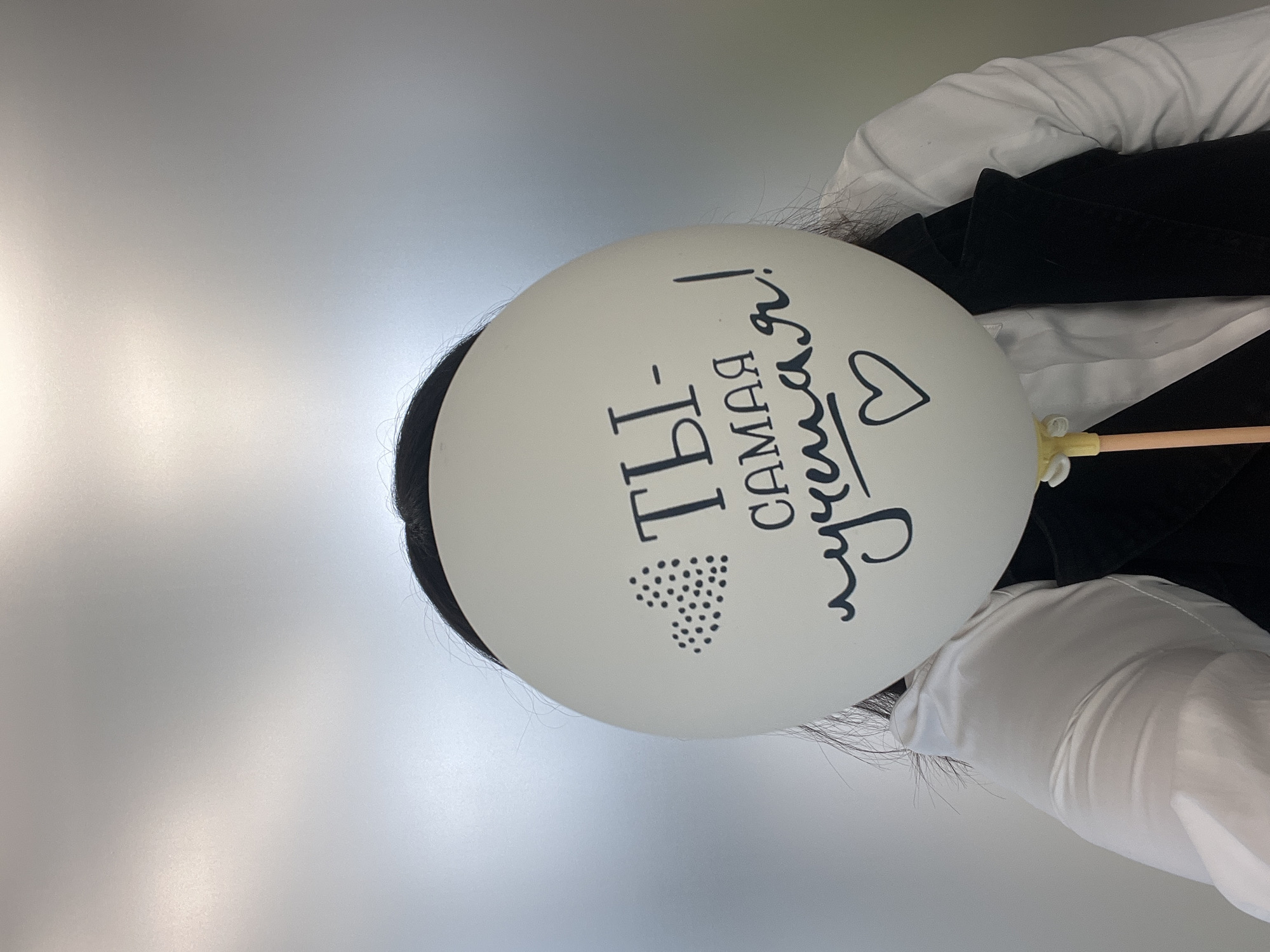 The image shows a person holding a white balloon with Cyrillic text that translates to "You are the best!" in English. The person is wearing a white collared shirt and a black jacket or blazer. Part of their dark hair is visible behind the balloon. The background is a blurry gradient of white and grey. The location, Perm, Russia, is not identifiable from the image.