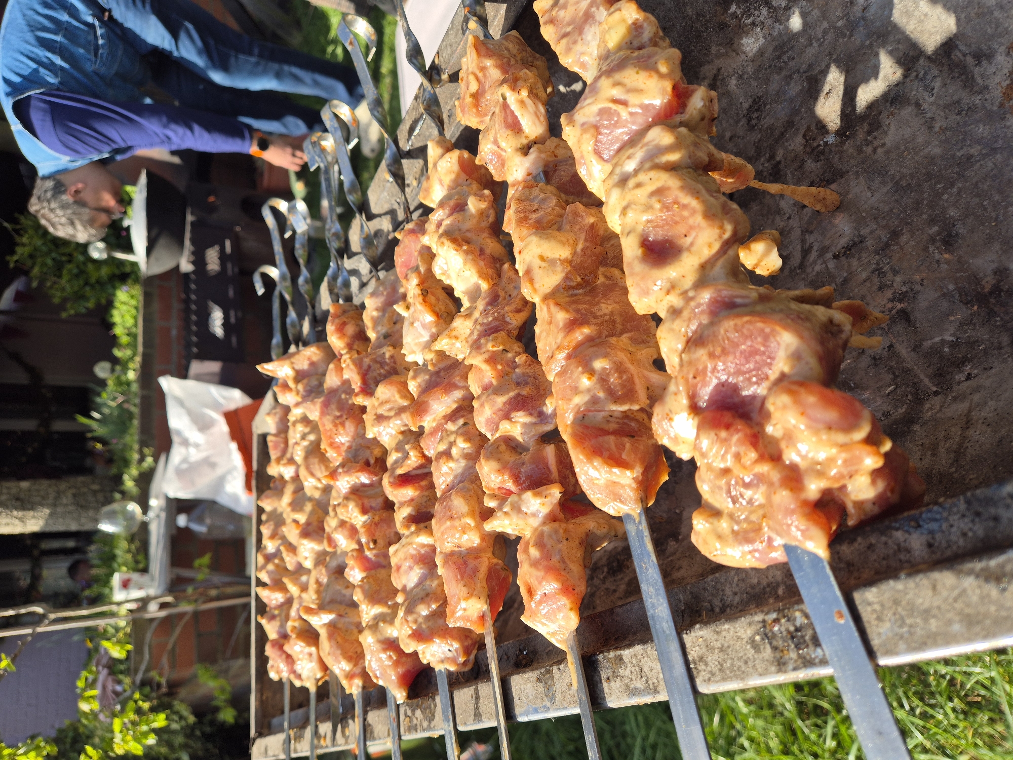 The image shows a close-up view of several skewers of marinated meat, likely pork or chicken, arranged on a metal barbecue grill. The meat is cut into bite-sized pieces and coated in a light-colored marinade. The skewers have twisted handles. A man is visible in the background, wearing a blue shirt and jeans. He is looking down, possibly attending to the grill. The background includes green grass, some foliage, a brick structure, and other indistinct items. This location is likely not in Ipswich, United Kingdom as the surrounding architecture does not align with that particular city. The scene suggests an outdoor barbecue or cookout is in progress.