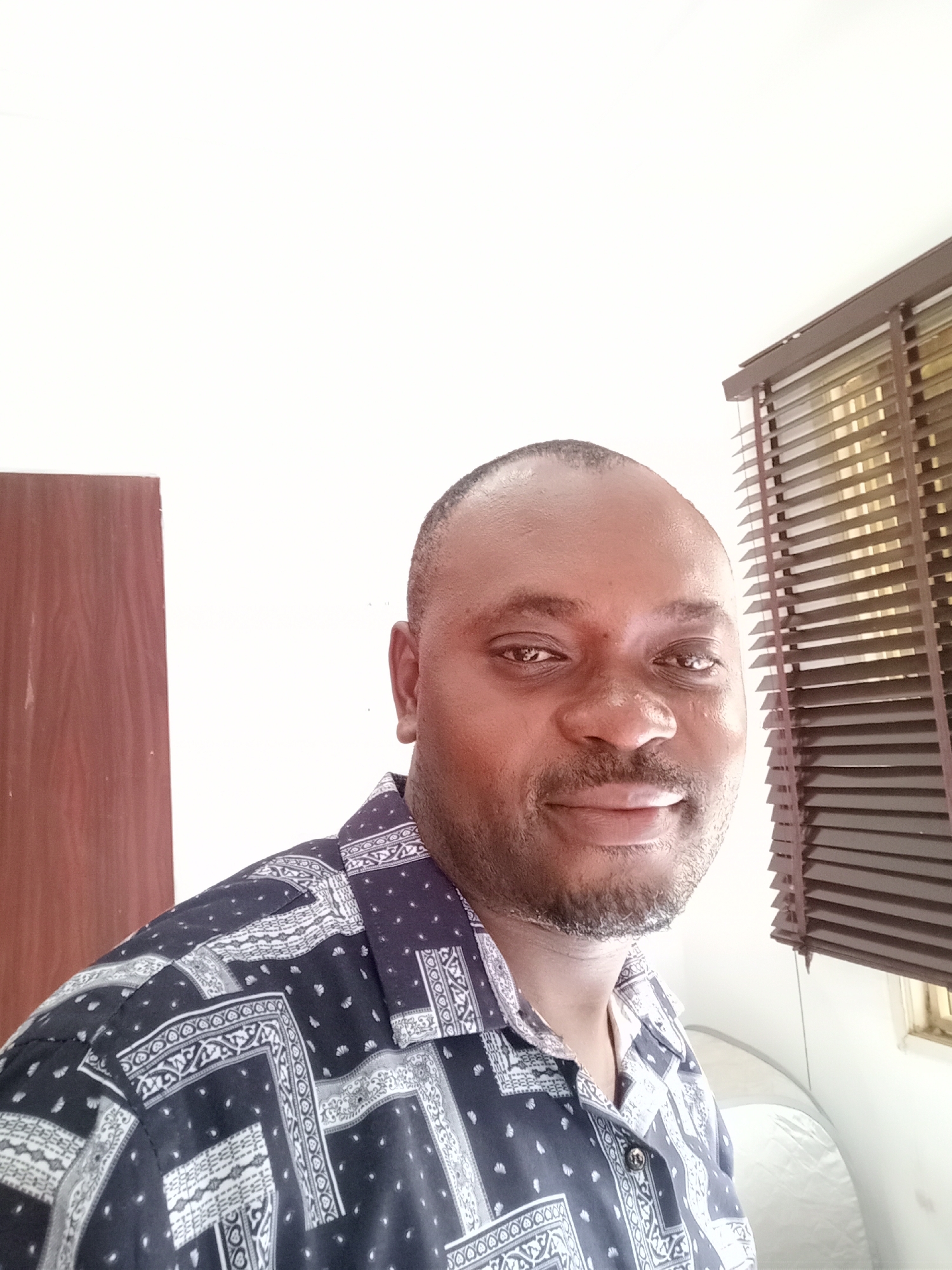 The image is a medium shot of a dark-skinned man, with a shaved head and a trimmed beard. He is smiling slightly and looking directly at the camera. He is wearing a patterned shirt with a dark blue base and white designs.
The man is indoors, standing in front of a white wall. To his left is a closed door. To his right, there is a window with closed brown blinds. The light source appears to be coming from the window.
Although the prompt mentions Gwarinpa, Nigeria, there is no visual information in the image that confirms the location. Therefore, the location cannot be verified from the image alone.