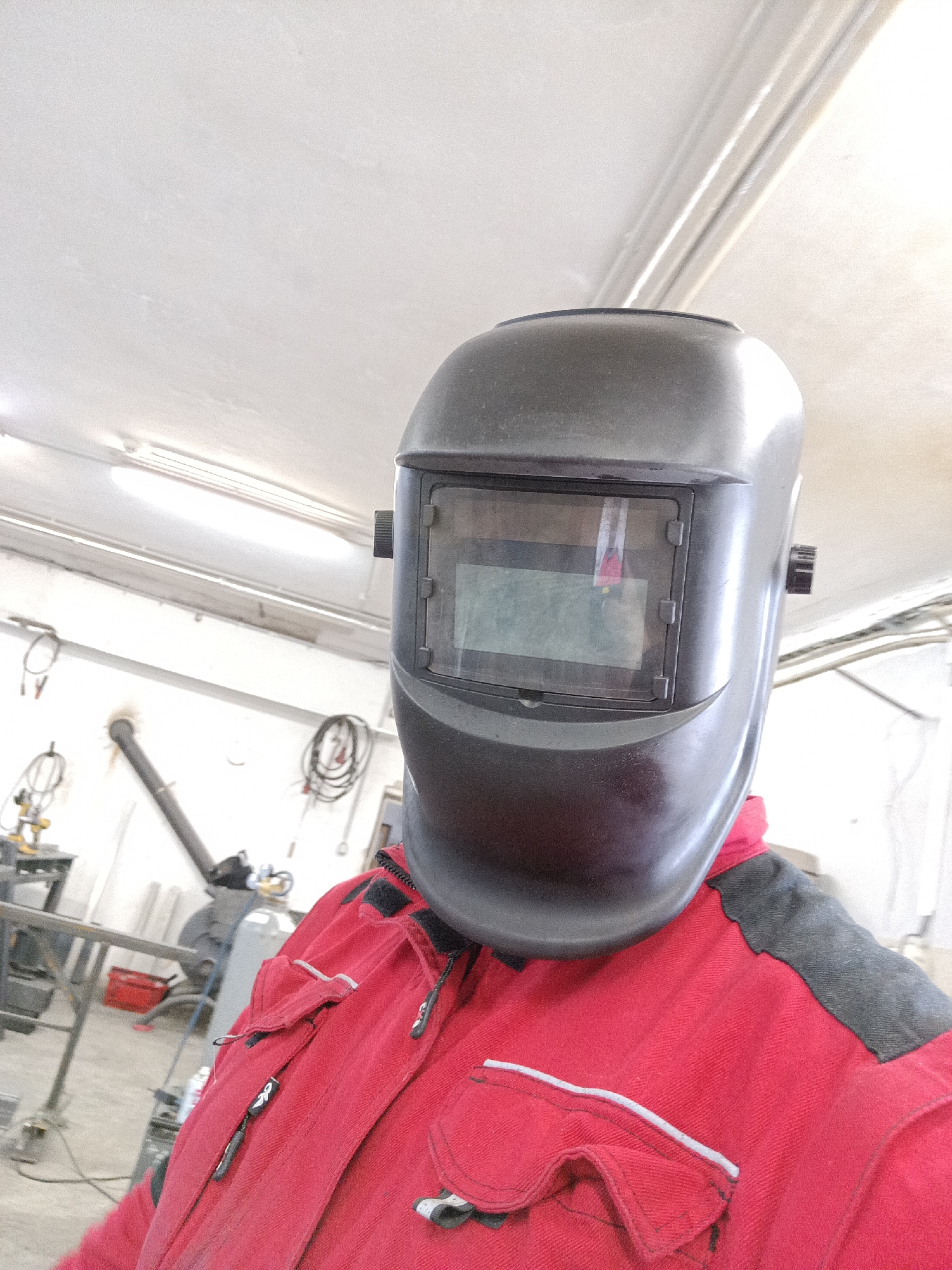 A close-up, waist-up selfie shows a person wearing a black welding mask and a red work jacket. The welding mask covers the entire face, with a rectangular viewing window that appears to be displaying some light. The jacket has several pockets with zippers and reflective trim. The background suggests an indoor workshop environment, with visible fluorescent lighting on the ceiling, pipes, wires, and industrial equipment such as a welding machine and a gas cylinder. The overall scene is dimly lit, with the exception of the bright ceiling lights. The location is identified as Dalovy, Czechia, indicating an industrial or manufacturing setting within that region.