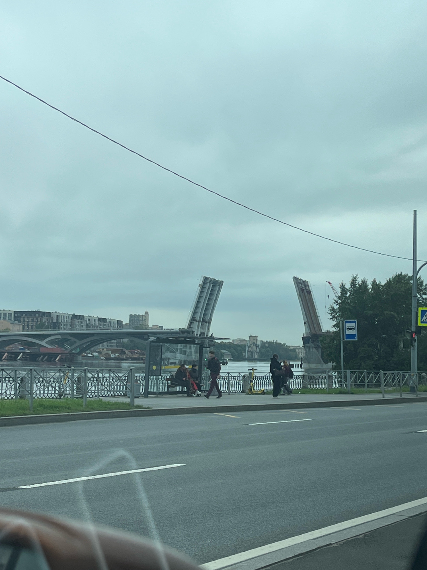 The image depicts a view of the Neva River in Saint Petersburg, Russia, on a cloudy day. A drawbridge is raised, with its two sections open to the sky, allowing passage for boats. In the foreground, a road with lane markings is visible, with a portion of a car's interior in the bottom left corner. Beside the road, there is a sidewalk with a bus stop shelter. Several people are seen on the sidewalk, with some sitting on a bench at the bus stop and others walking. A yellow scooter is parked near the bus stop. A decorative metal fence separates the sidewalk from the river. In the background, buildings line the opposite bank of the river, and trees are visible on the right side of the frame. A traffic light and a pedestrian crossing sign are also present. The overall scene suggests a typical urban environment with a functioning drawbridge and public transportation.