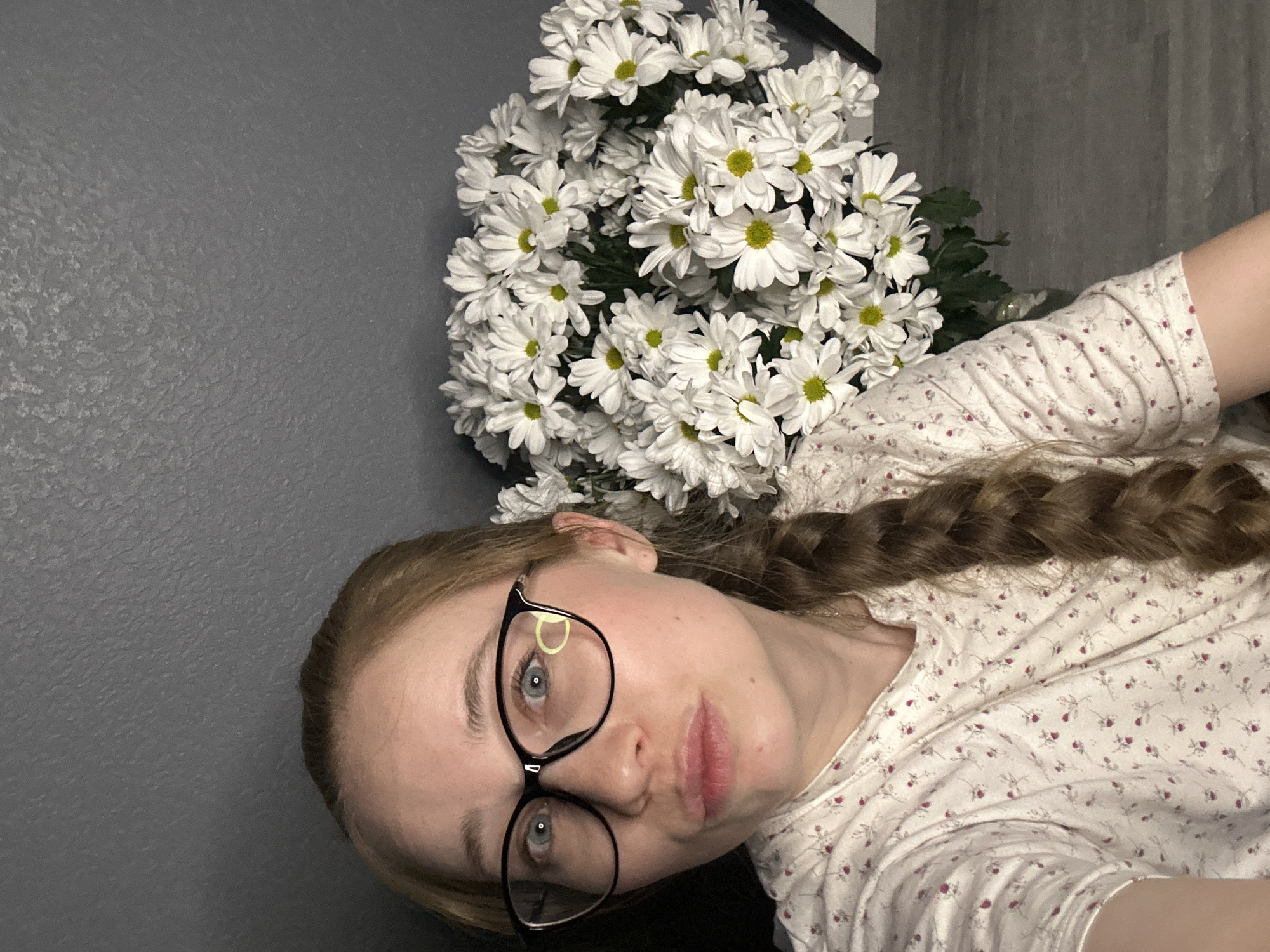 A woman with light skin, wearing black-rimmed glasses and a white floral-patterned shirt, poses for a selfie. Her hair is light brown and braided. To her right is a large bouquet of white daisies with yellow centers. The background consists of a dark gray wall. There is no indication of Saint Petersburg, Russia.