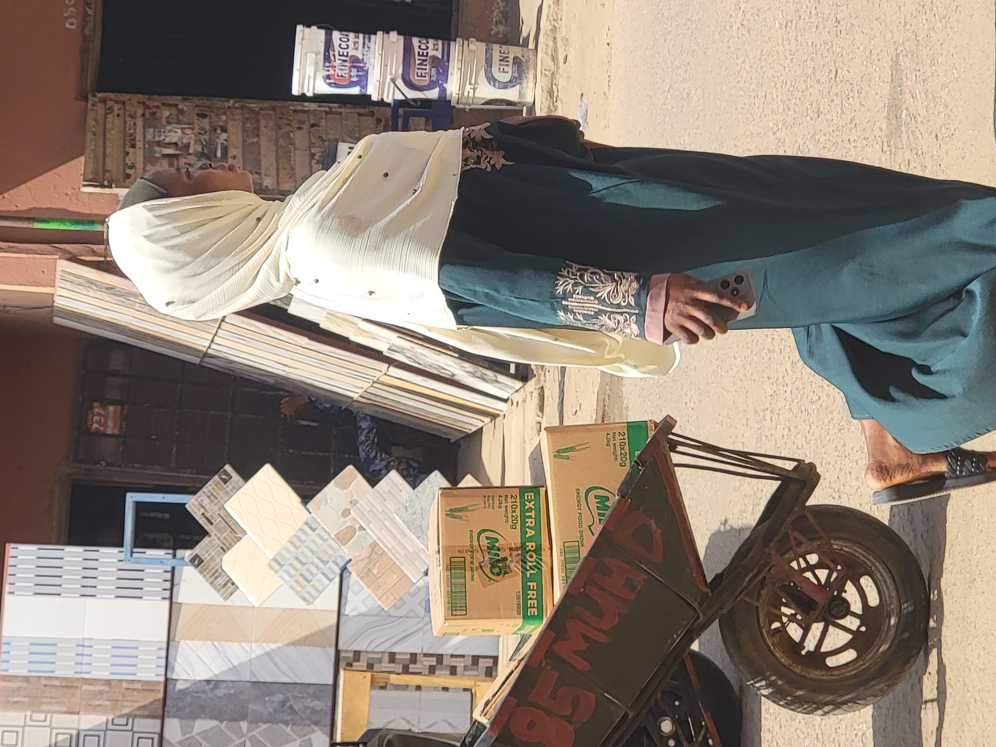 A woman wearing a green dress and white headscarf stands on a sunny street, holding a mobile phone. Behind her are stacks of ceramic tiles and containers labeled "FINECO." A wheelbarrow, partially obscured, is loaded with boxes of "Milo Energy Food Drink" marked "EXTRA ROLL FREE," with the text "85 MUHD" spray-painted on it. Her feet are visible, clad in sandals. The scene is likely a commercial area, possibly in Maiburiji, Nigeria.