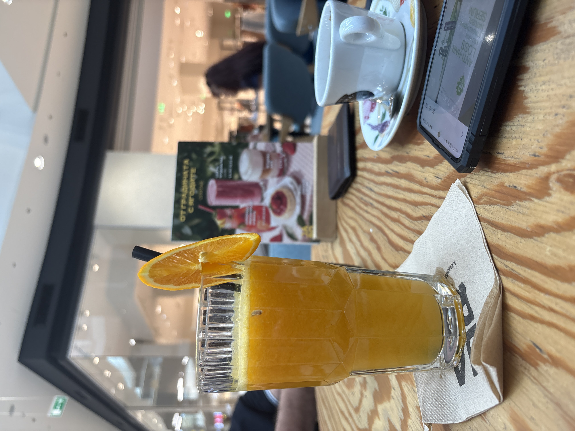 The image shows a glass of orange juice with a slice of orange on the rim, placed on a napkin on a wooden table. Next to the juice are a teacup on a saucer and a phone. In the background, a book with images of fruits and drinks is visible, and further in the background, a blurred figure of a person sitting in a cafe setting can be seen, illuminated by the lights from the window. The phone screen shows "ANTI-HAIR LOSS" and other text, indicating an advertisement. The text on the book cover reads "с ягодите от градината", which translates to "from the garden with berries" indicating it might be a menu or catalog for smoothies and juices. The napkin appears to have the word "LEMON" printed on it. Based on the text on the menu, the photo was most likely taken in Burgas, Bulgaria.